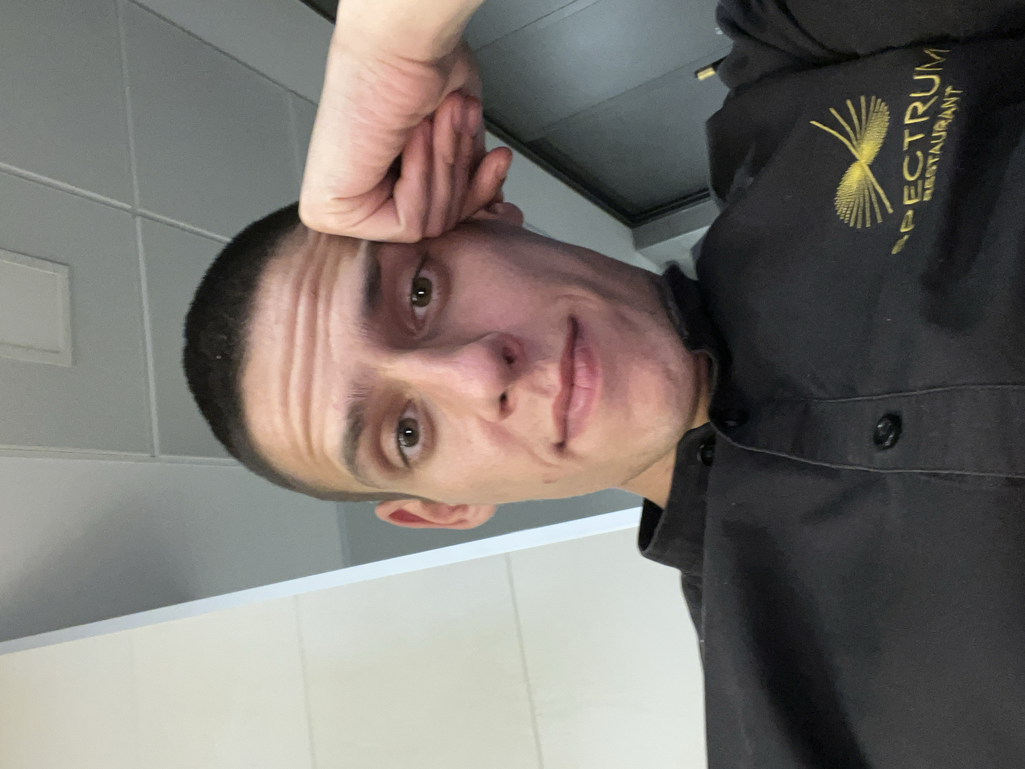A portrait-oriented, eye-level shot captures a young man from the chest up, looking directly at the camera with a neutral expression. He has short, dark hair and brown eyes. His right hand is positioned on the side of his head, with fingers bent, suggesting a moment of thought or weariness. He is wearing a black collared shirt, likely a uniform. Prominently displayed on the left side of his shirt is a gold embroidered logo that reads "SPECTRUM RESTAURANT", accompanied by a stylized fan or sunburst graphic above the text.
The background consists of light grey tiled walls, with a distinct white horizontal surface visible in the lower portion of the frame, possibly a counter or partition. The indoor setting, with its tiled surfaces and uniform, suggests a professional environment such as a restaurant kitchen or service area. The lighting is consistent and even, typical of artificial indoor illumination. The overall image appears to be a straightforward snapshot, possibly a selfie. While the context states the location is Sofia, Bulgaria, this information is not discernible from the visual cues within the image itself.