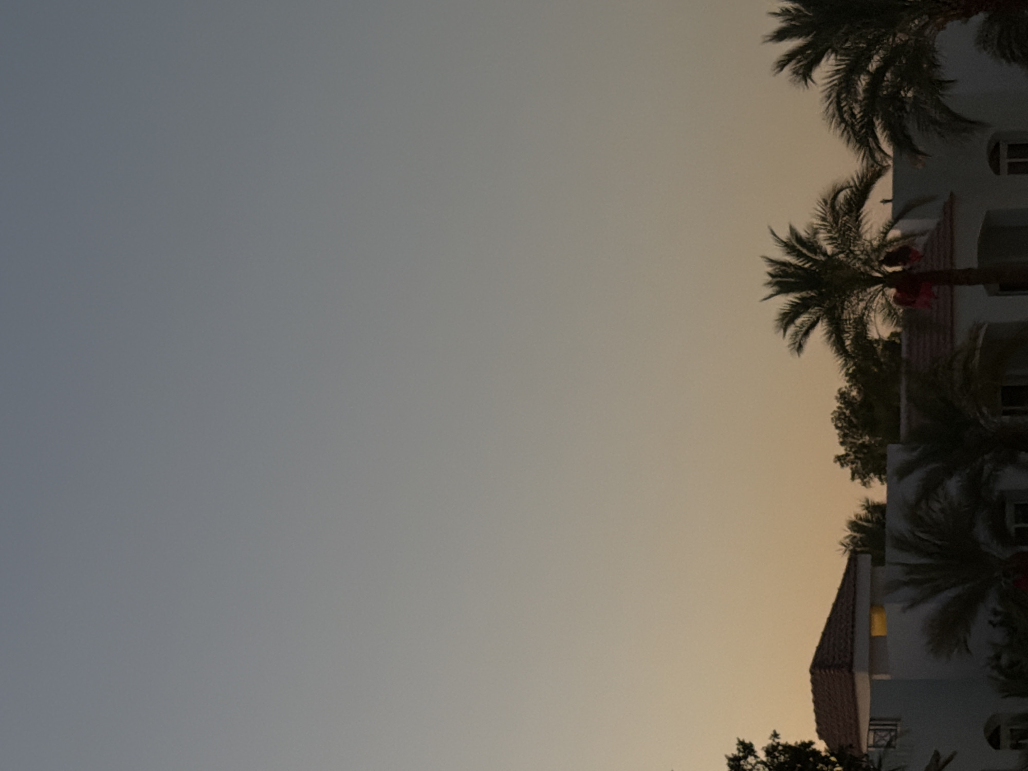 The image shows the twilight sky over a resort area in Sharm El Sheikh, Egypt. The sky transitions from a pale blue at the top to a warm orange hue closer to the horizon. Silhouetted against the sky are the tops of buildings with tiled roofs and palm trees. One building has a light on inside, illuminating a section of the roof. The scene appears to be from a tropical or subtropical location given the presence of palm trees.