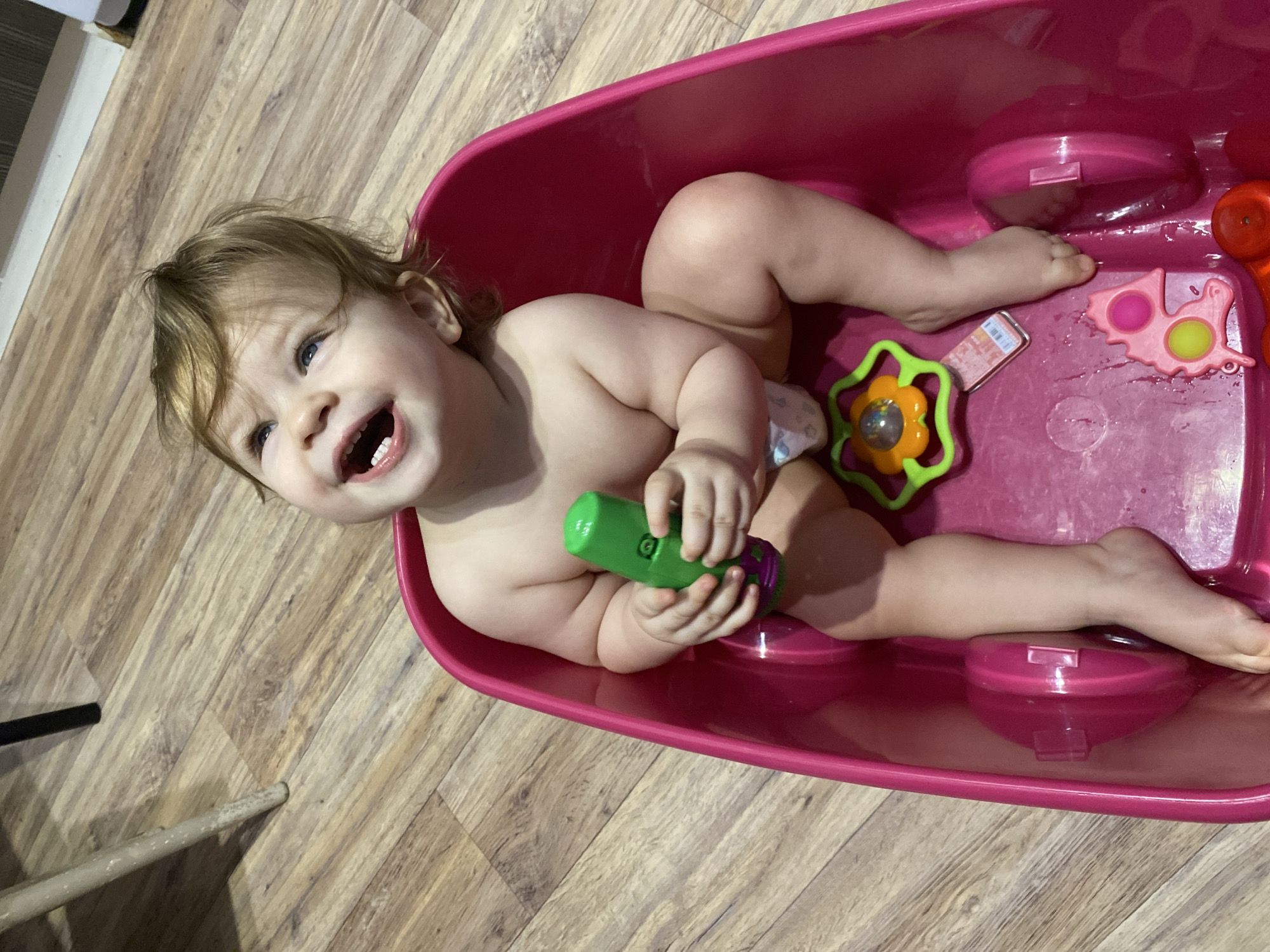 A toddler sits in a pink bathtub filled with water and toys on a wood floor. The child is smiling and holding a green and purple toy. The bathtub also contains a green flower-shaped toy, a pink butterfly-shaped toy, and a red toy. The location appears to be inside a residential home.