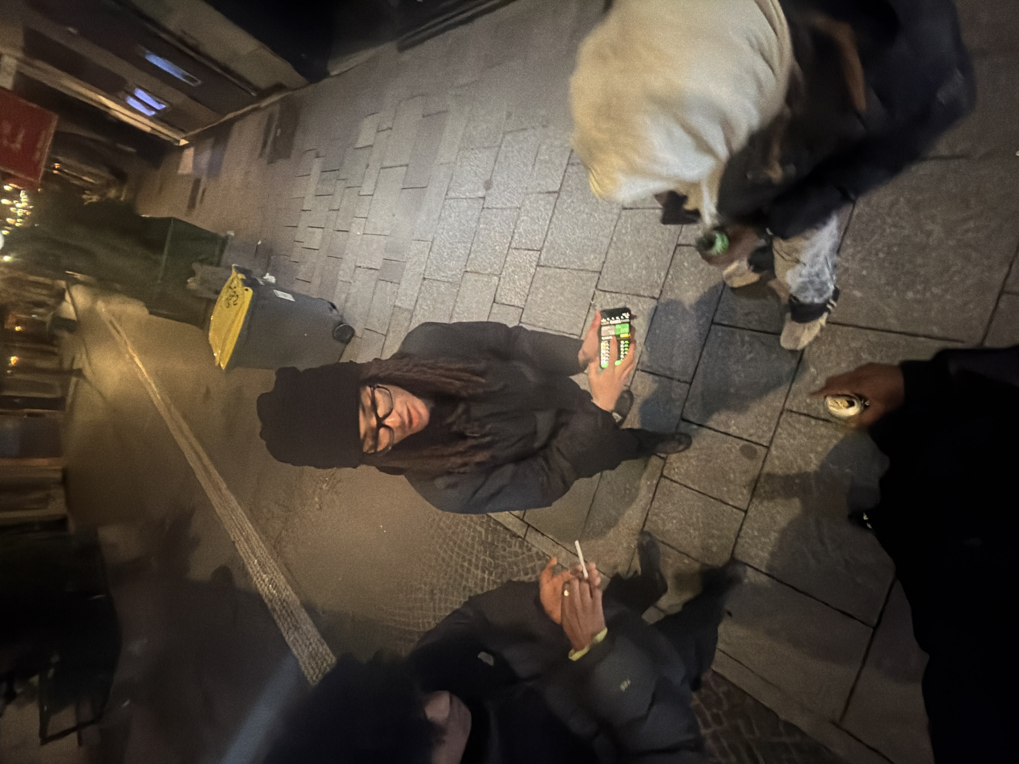 The scene depicts three individuals gathered on a paved urban sidewalk at night in Paris, France. A central figure, wearing a black beanie, black-rimmed glasses, and dark clothing, stands and holds a smartphone horizontally with both hands, its screen illuminated and displaying a colorful, grid-like interface. To the left, a second individual, partially visible and wearing a dark puffy jacket, holds a lit cigarette in their right hand, with a yellow band on their left wrist. To the right, a third individual, partially visible and wearing a light-colored top and dark pants, is seated or crouching on the sidewalk, holding a silver can in their right hand. A furry, light-colored object rests near this individual's legs. The background features a dark asphalt street with a faint white line, illuminated building facades, and several waste bins, including two large green bins and one gray bin with a yellow lid, positioned along the sidewalk.