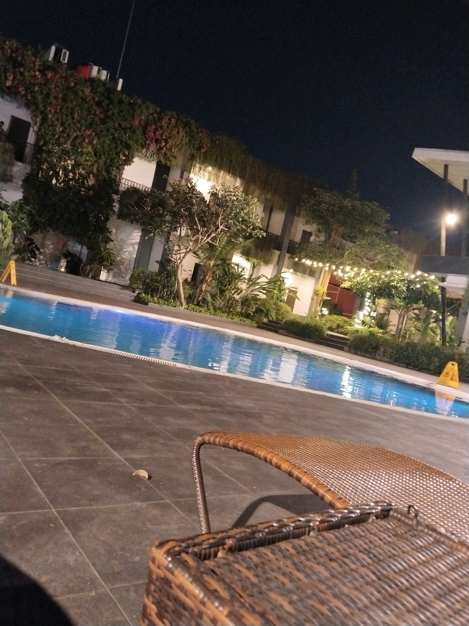 An exterior night-time, low-angle shot shows a swimming pool in front of a multi-story building. The building is covered in green vines and illuminated by string lights. The pool is lit from below, creating a blue glow in the water. A wicker lounge chair is in the foreground, partially obscuring the view of the pool and the building. The scene is likely in Depok, Indonesia, a city known for its tropical climate and lush vegetation, making it a plausible setting for an outdoor pool area with abundant greenery. The image captures a serene and inviting atmosphere, suggesting a resort or hotel setting.