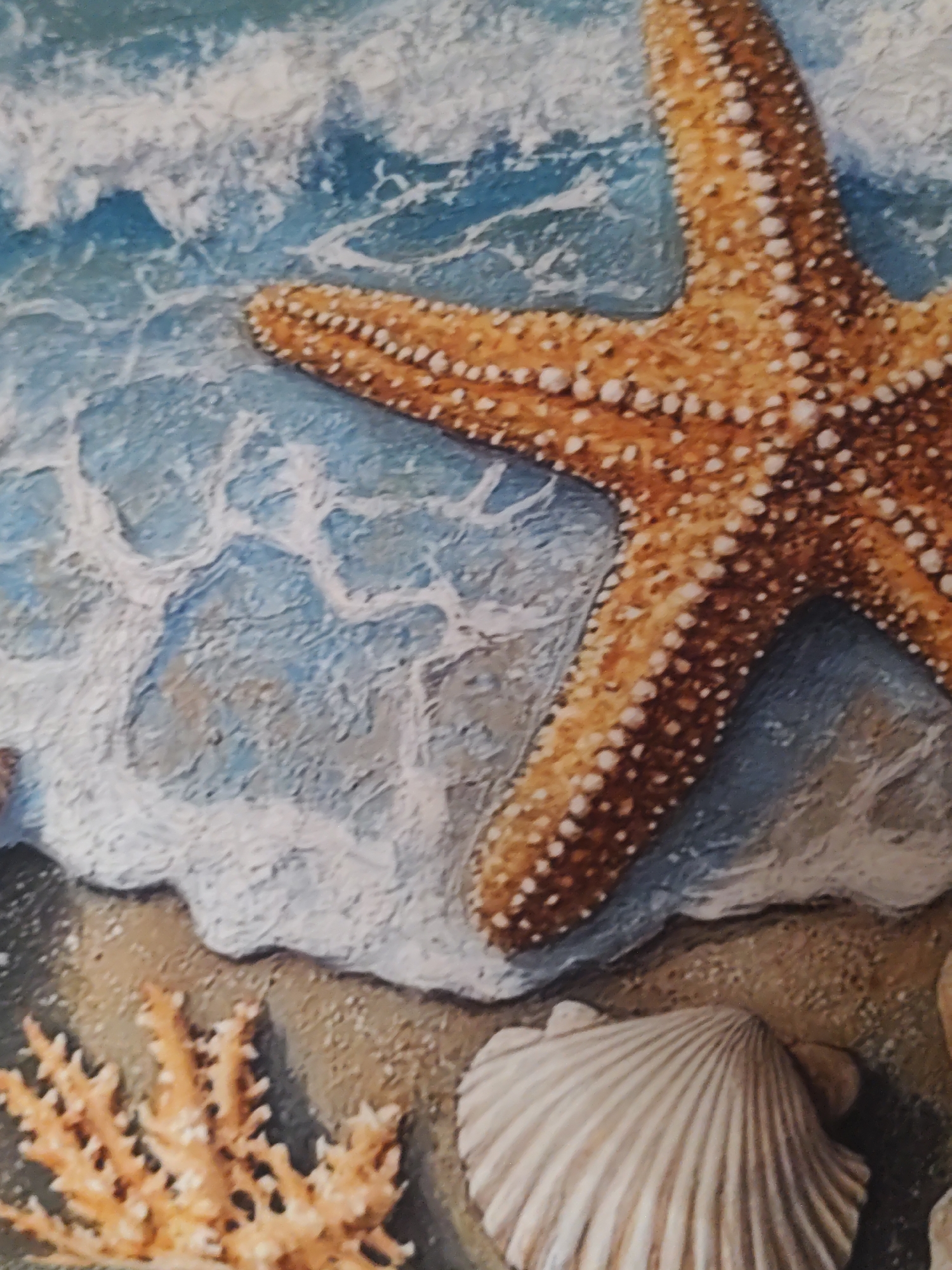 The image depicts a close-up view of a textured artwork featuring a starfish resting on a sandy seabed with incoming waves. The starfish is orange and speckled with white. The water is depicted with blue hues and white frothy foam, creating a sense of movement. In the lower left corner, there is a piece of light orange coral, and in the lower right corner, a beige scallop shell is visible. The overall impression is a stylized marine scene, likely a decorative piece rather than a photograph of a real-world location. There is no information in the image to indicate its location in Montana, Bulgaria.
