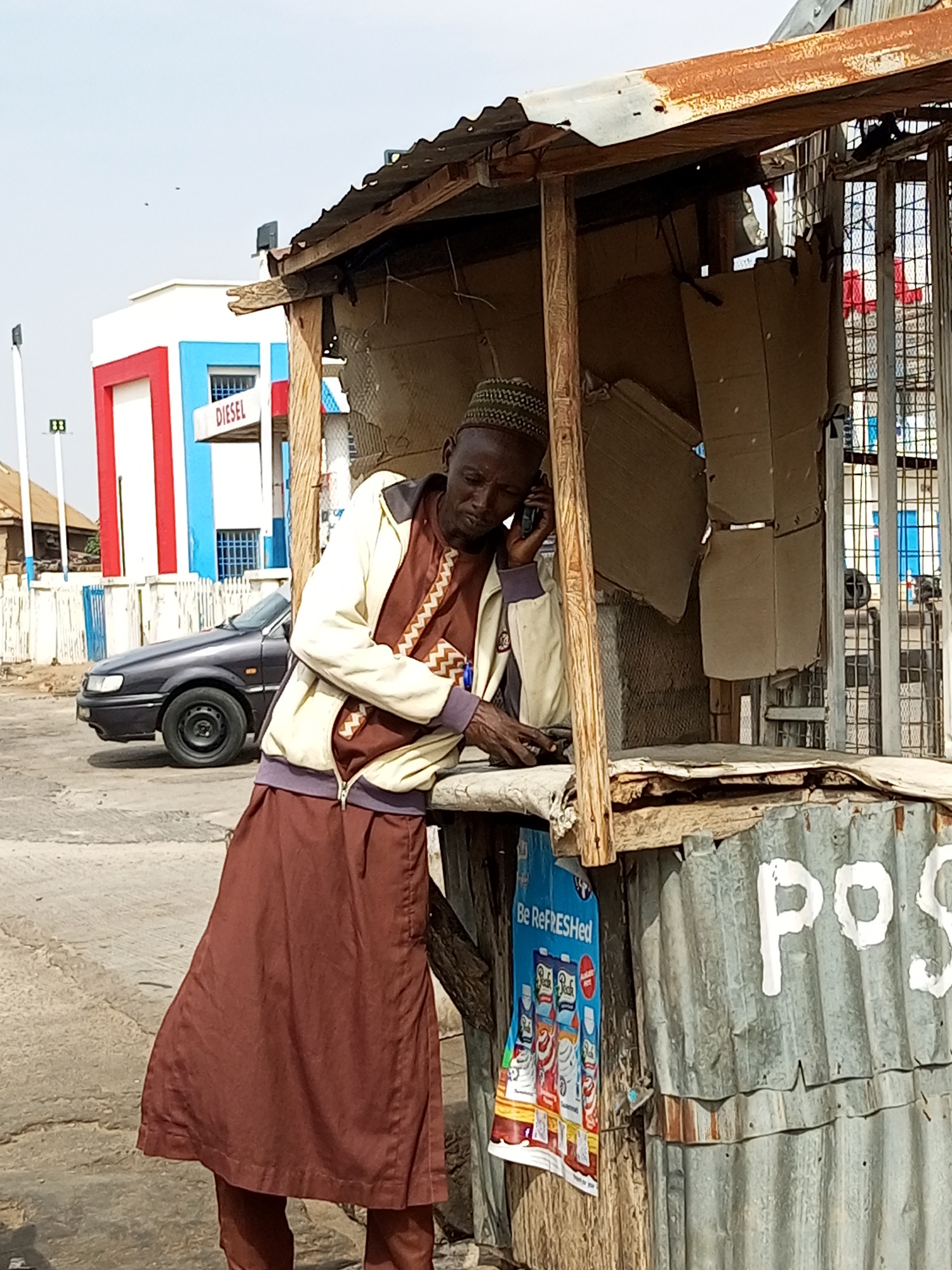 The image depicts a man in Gombe, Nigeria, standing at a makeshift kiosk. He is wearing a patterned kufi cap, a beige jacket with a brown and white patterned shirt underneath, and a brown skirt over trousers. He is holding a mobile phone to his ear and appears to be engaged in a conversation. The kiosk is constructed from corrugated metal and wood, with cardboard panels visible inside. A sign with the word "POS" is painted in white on the corrugated metal exterior. A blue poster with the text "Be ReFRESHed" and images of beverage cartons is attached to the wooden side of the kiosk. In the background, a dark-colored sedan is parked near a building with red and blue vertical stripes and the word "DIESEL" visible. The sky is clear and overcast. The scene suggests a street-side vendor or service point in an urban environment.