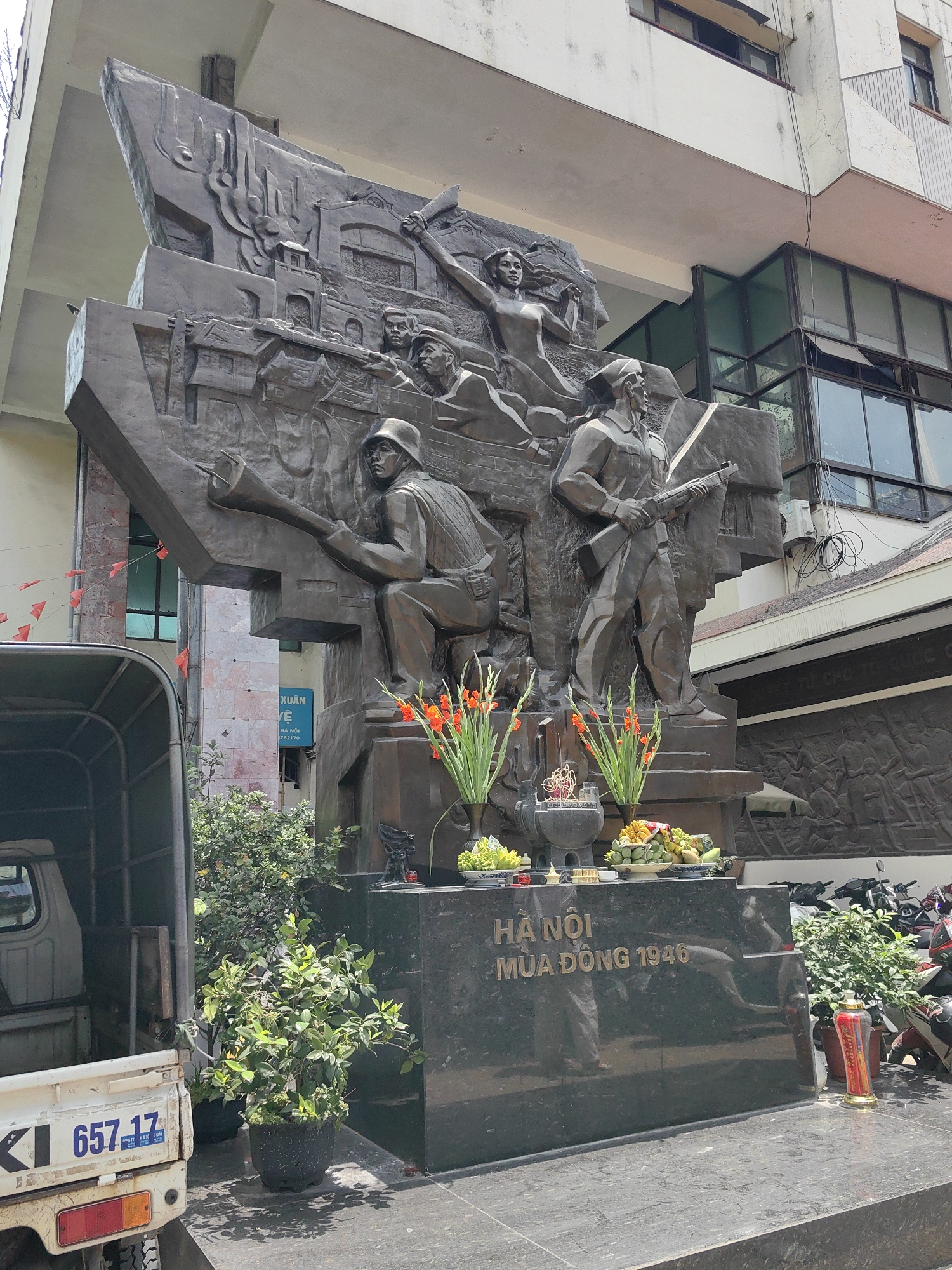 A large, dark bronze monument commemorating "HÀ NỘI MÙA ĐÔNG 1946" (Hanoi Winter 1946) stands prominently in an urban street setting in Hà Nội, Vietnam. The monument features multiple heroic-style figures in action, including a soldier in a helmet kneeling with a large weapon, another standing with a rifle, and a dynamic female figure with outstretched arms. The background of the sculpture depicts architectural elements suggesting urban combat.
The monument rests on a multi-tiered black marble base, where offerings of fresh green and yellow fruits, vibrant orange gladioli flowers, and incense are carefully placed in dark vases and bowls, indicating ongoing reverence. Potted green plants are arranged around the base.
Behind the monument is a multi-story, light-colored building with windows and balconies, showing signs of wear. On the left, the rear of a white truck (Suzuki brand) with license plate "657.17" is visible. Small red triangular flags are strung across the street to the left. In the background to the right, motorbikes are parked, and a textured relief wall is partially visible with the text "QUYẾT TỬ CHO TỔ QUỐC CŨ" (Determined to Die for the Fatherland) in relief. A blue sign in the background reads "XUÂN VÊ" (Spring Returns/Spring is coming) with a phone number "282170".
The scene is brightly lit by daylight, suggesting a clear day. The monument commemorates a pivotal historical event, depicting the struggle and sacrifice during the First Indochina War.