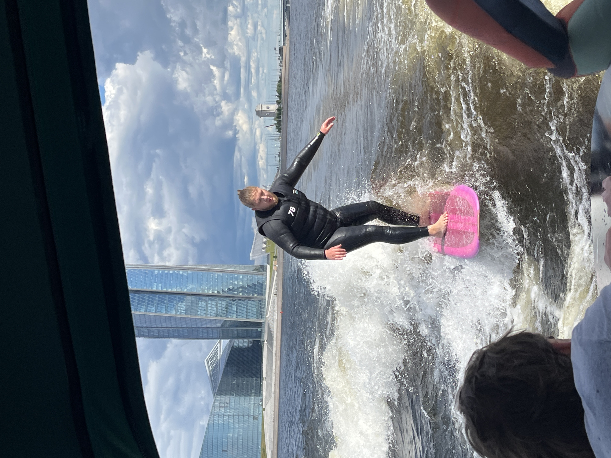 A man is wakeboarding on the Neva River in Saint Petersburg, Russia, with the Lakhta Center skyscraper and its adjacent complex in the background. The man is wearing a black wetsuit with a life vest and is standing on a pink wakeboard. He is creating a large spray of water as he rides. In the foreground, the back of a person's head is visible, suggesting they are in a boat. The sky is partly cloudy with dramatic cloud formations. The architecture in the background is modern and striking, characterized by glass facades and a tall, slender skyscraper. The Neva River is wide and dark, reflecting the sky. The overall scene captures an active moment against a prominent urban backdrop.