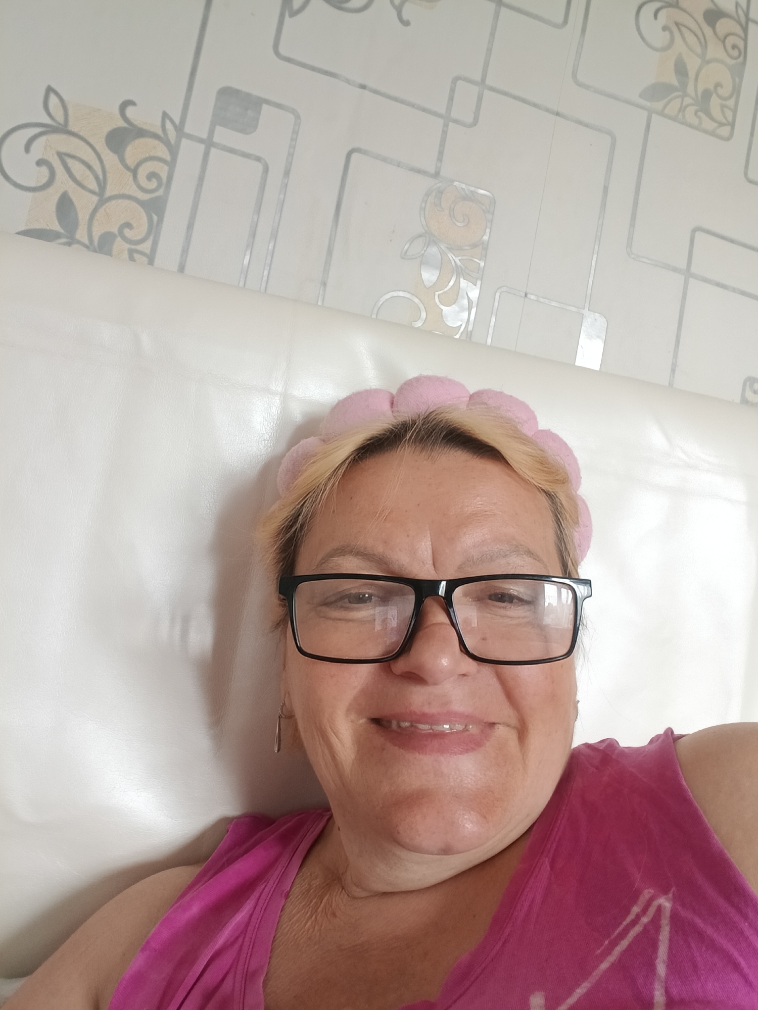A woman is in a room in the city of Красный Маяк, Russia. She is wearing a pink top, glasses, and a pink head band. The background includes a white headboard and patterned wallpaper. She is smiling and looking directly at the camera.