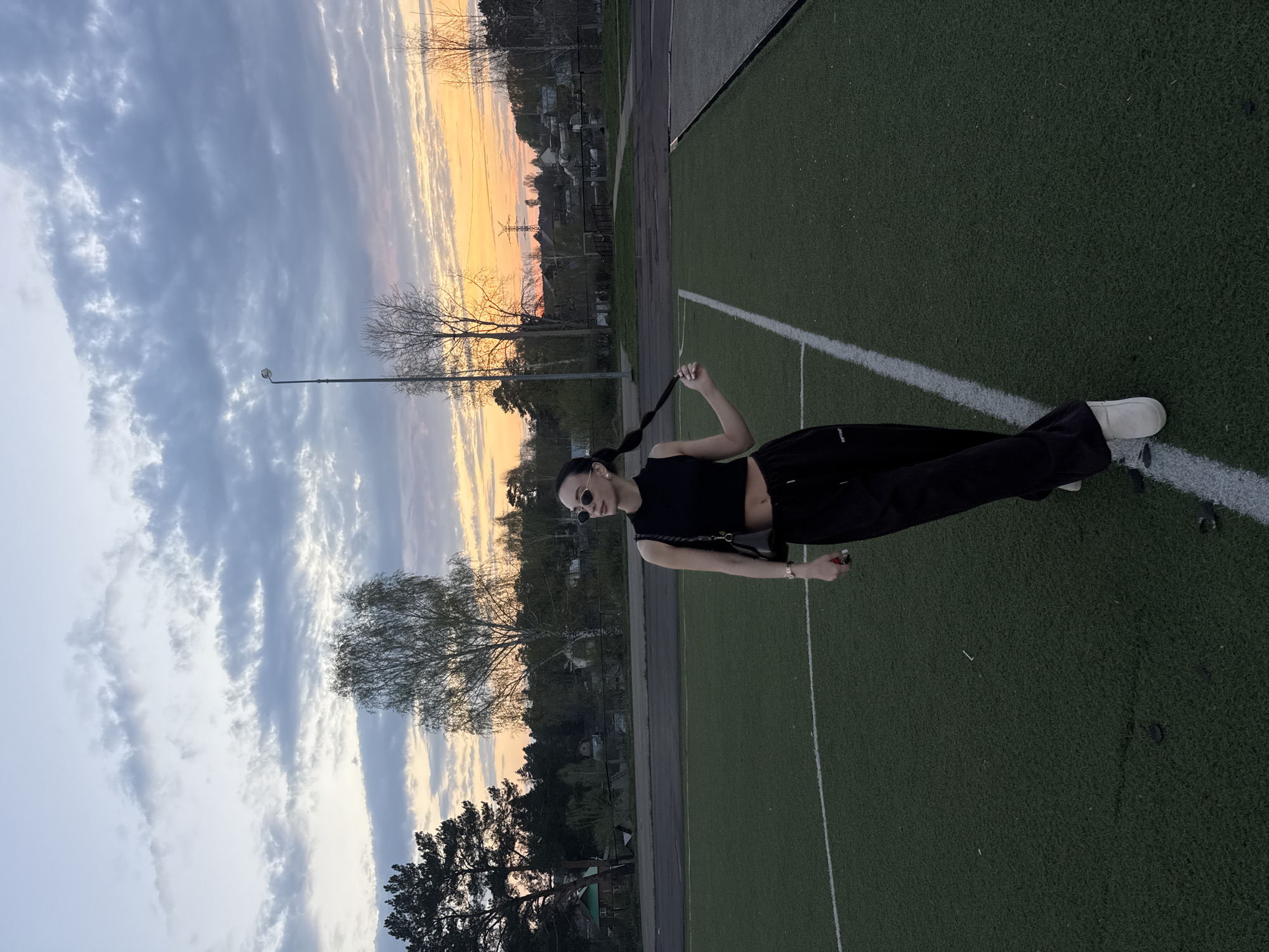 A young woman stands on an artificial turf field with white boundary lines. She wears black clothing, sunglasses, and white shoes. She holds the end of her braid. The background includes a cloudy sky with a sunset glow, trees, and distant buildings. The scene is in the city of Chaikovsky, Russia.