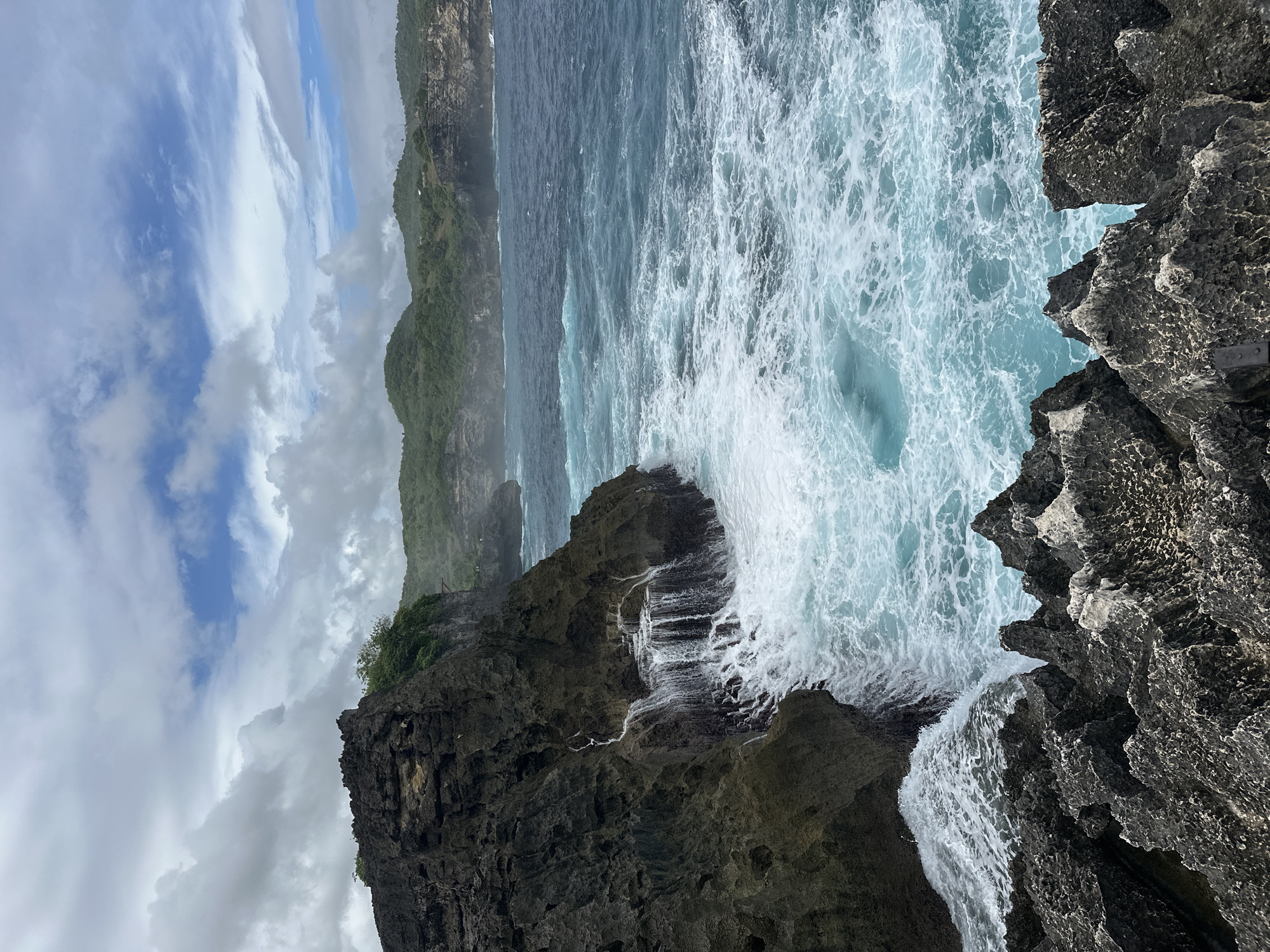 The image captures a coastal scene in Nusa Penida, Indonesia. Rugged, dark gray rock formations dominate the foreground, meeting the turbulent turquoise water of the ocean. The water is churned with white foam, indicating strong wave action. In the distance, a green, hilly coastline stretches under a partly cloudy sky. The sky displays a mix of blue and gray clouds.