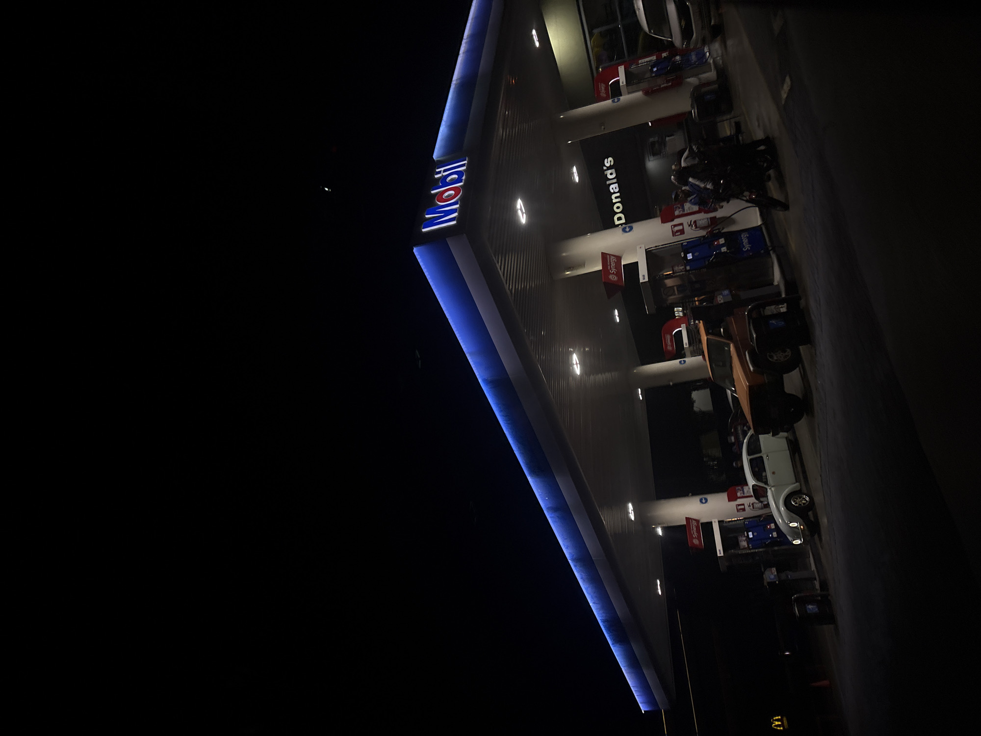 The image shows a Mobil gas station with a McDonald's in San Andrés Cholula, Mexico at night. Several cars are parked at the gas pumps. The Mobil sign is illuminated with blue lighting.