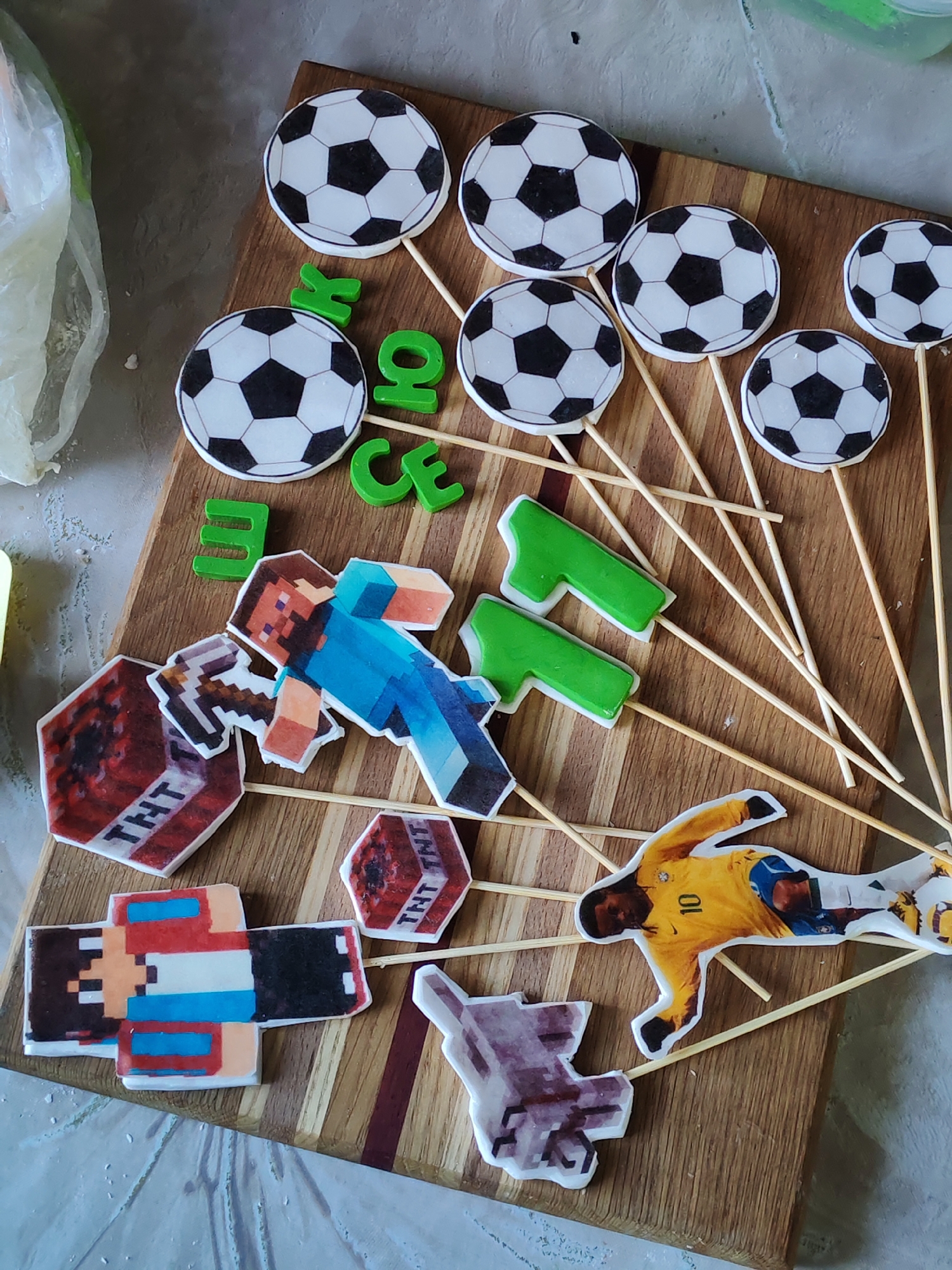 The image shows a wooden cutting board with various decorative elements. These include:
- Several soccer ball decorations on sticks.
- Green plastic letters spelling out a name, possibly "BRYCE".
- Minecraft-themed decorations like "TNT" blocks, a character resembling a "Steve" figure, and other pixelated designs.
- A soccer player cutout wearing a yellow jersey with the number 10.
The elements are arranged randomly on the board, suggesting they might be intended for a cake or other celebratory display. The background is blurred, showing a glimpse of a white surface and a translucent plastic bag.