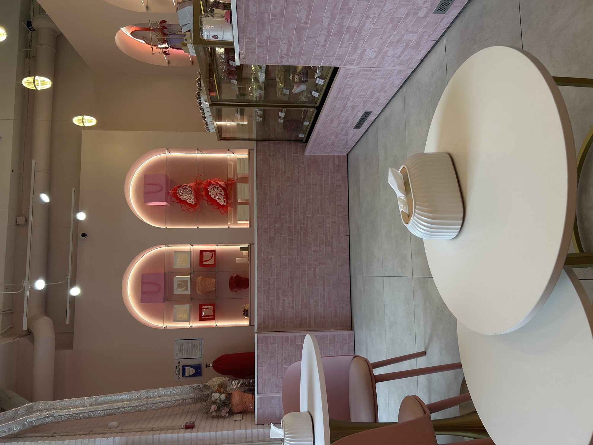 The image depicts the interior of a confectionery or bakery with a pastel pink color scheme. The establishment features a display counter with glass cases filled with desserts, likely cakes and pastries. Behind the counter, shelves are arranged within arched displays, showcasing decorative items such as gift bags, floral arrangements, and vases. Seating areas are visible with round white tables and pink chairs. One table in the foreground has a white tissue box. The walls are tiled with a pink brick pattern, and the floor is tiled with large square gray tiles. Overhead, industrial lighting fixtures and ventilation pipes are present. The environment appears clean and well-lit, suggesting it is open for business. No people are visible in the image. The overall aesthetic is modern and feminine.