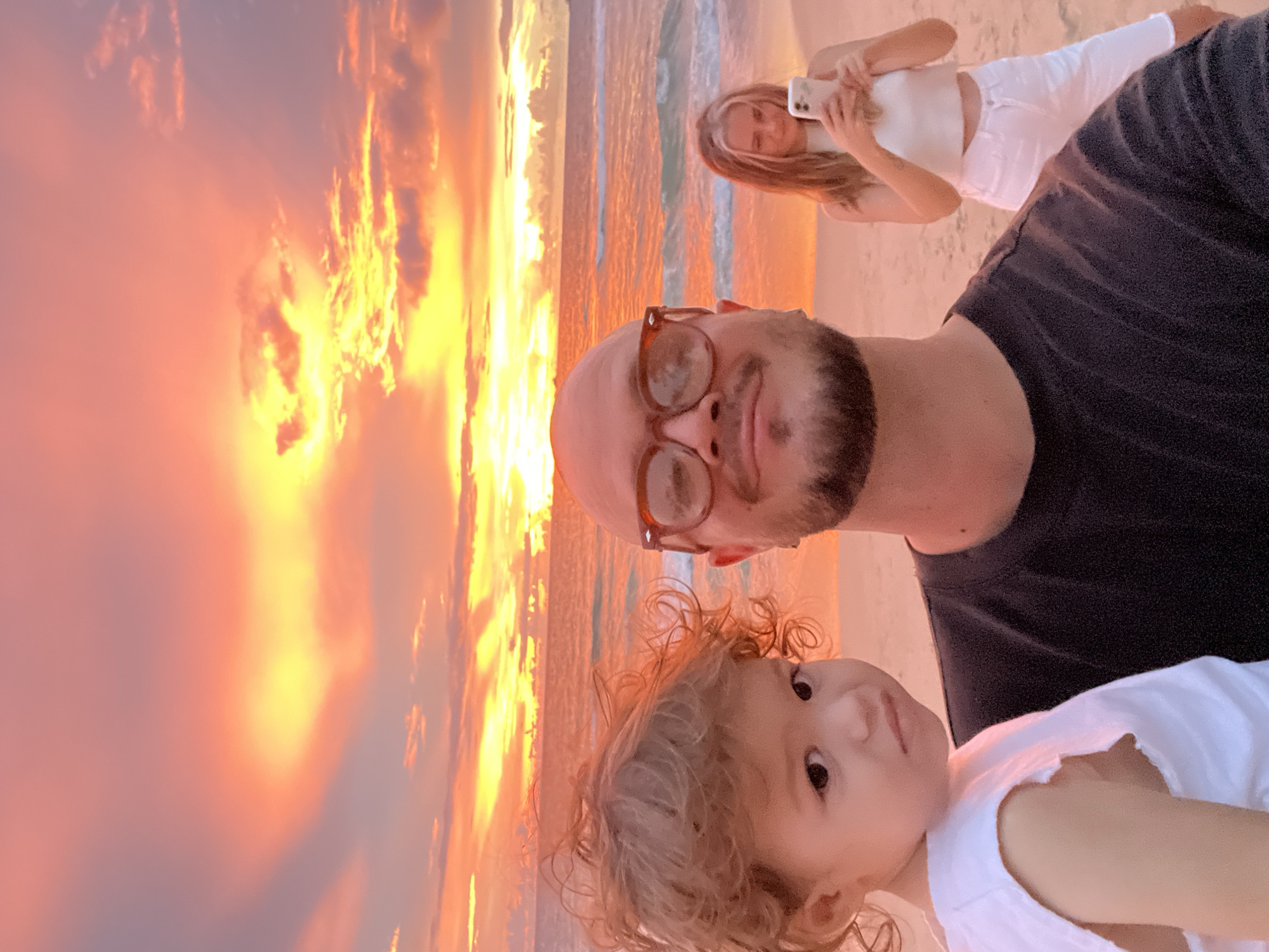 A selfie taken by a man with a beard and glasses shows him holding a young child, with a woman in the background. They are on a beach at sunset, with the sky filled with vibrant orange and yellow hues. The ocean is visible behind them, with gentle waves rolling onto the shore. The man is wearing a black t-shirt, and the child is wearing a white top. The woman in the background is wearing a white crop top and white pants, and she is holding a smartphone, seemingly taking a picture. The scene is bathed in the warm glow of the setting sun.