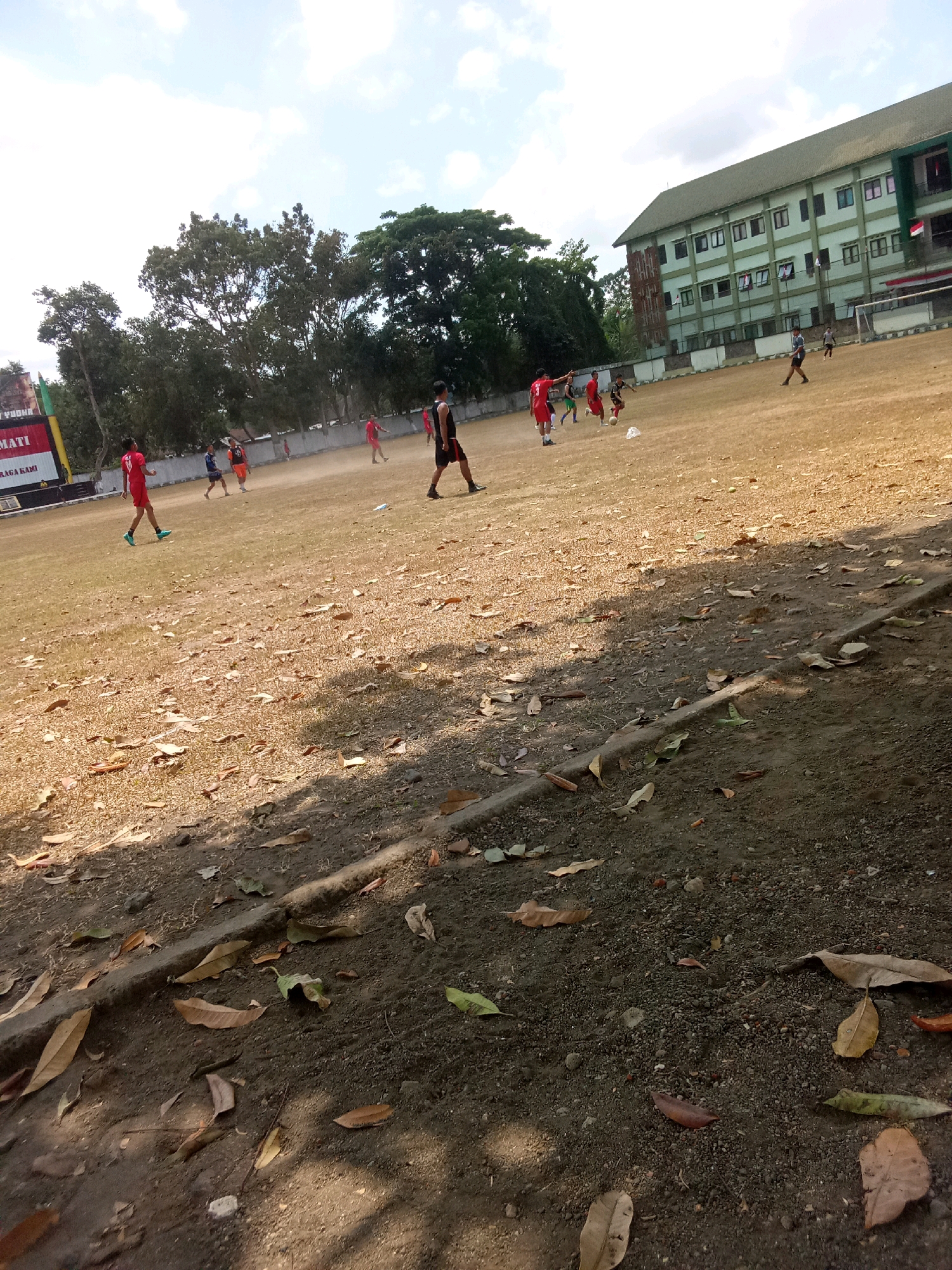 The image depicts a soccer field in Kebonsari, Indonesia, with several young men engaged in a game. The field is dry and dusty, with patches of sparse grass. A large, multi-story building with a green roof and white walls stands in the background, likely a school or dormitory. Some trees and a fence surround the field. The sky is partly cloudy. The overall impression is that of a casual game on a warm, sunny day.
