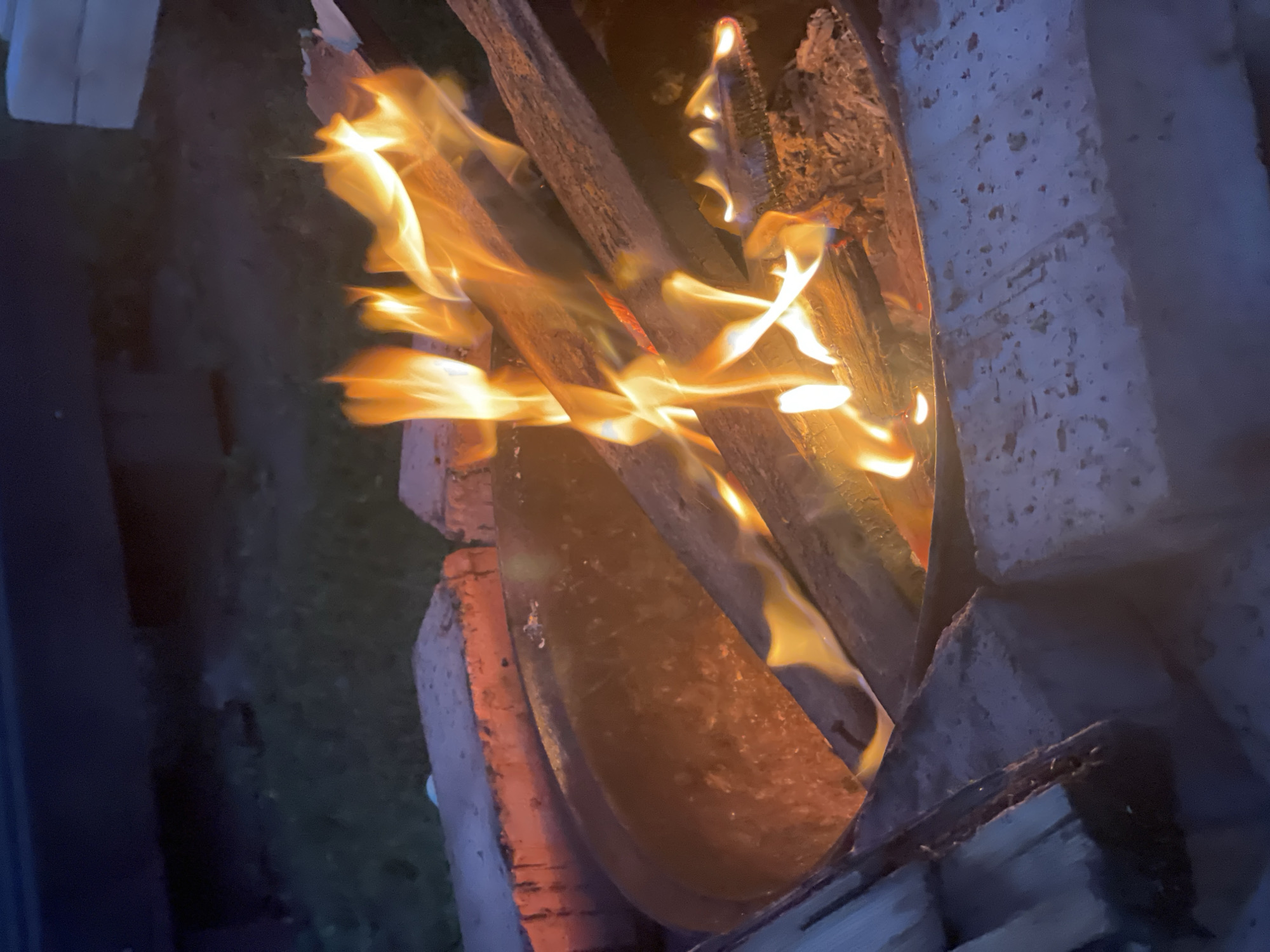 An outdoor fire pit is filled with burning wood. The fire is producing bright orange and yellow flames that dance above the wood. The fire pit itself is made of dark metal and is surrounded by bricks. The scene is dimly lit, suggesting it is either dusk or nighttime. The background is dark and out of focus, but appears to be an outdoor setting with some vegetation.