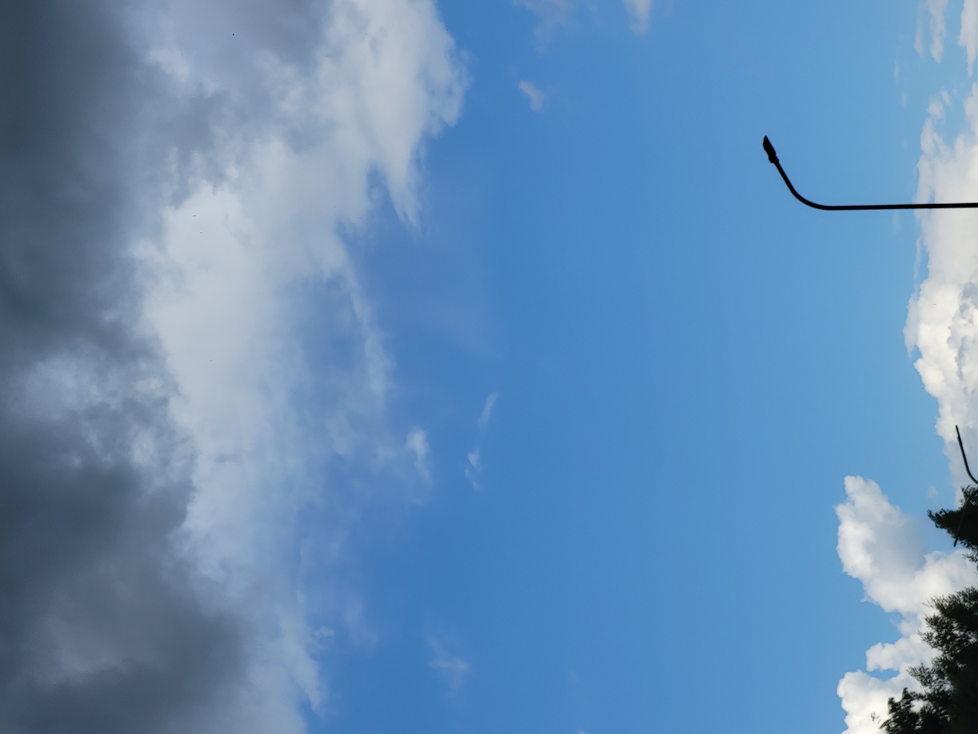 The image depicts a section of the sky with clouds and a silhouette of a street lamp. The sky is predominantly blue, with varying degrees of cloud cover. In the upper portion of the image, dark, heavy clouds are visible, suggesting the possibility of impending weather changes. Below these, lighter, white clouds are scattered across the blue sky. At the bottom of the frame, the dark silhouette of a street lamp is prominent, with its curved arm extending upwards and to the right. The base of the lamp is not visible. To the lower left of the frame, the dark silhouettes of trees are partially visible. The scene appears to be an outdoor environment, possibly a street or park, in Ozolnieki, Latvia. There are no people or any explicit actions or interactions taking place. The overall impression is one of a natural landscape with man-made elements.