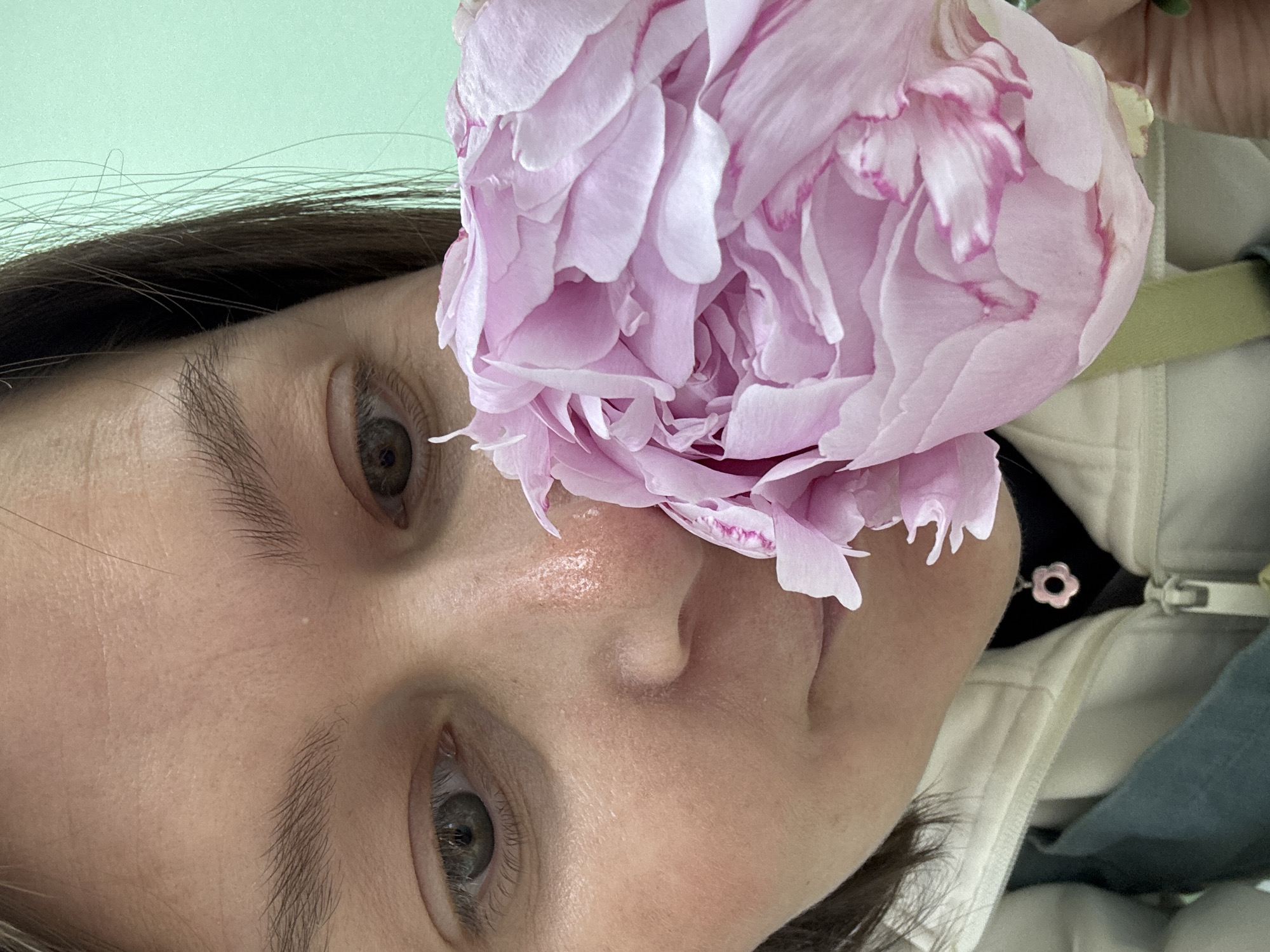 A close-up image captures a person's face partially obscured by a large, light pink peony. The individual has fair skin, dark brown hair, and light-colored eyes. The peony is positioned near the nose and upper cheek of the person. A white jacket with a dark neckline is partially visible. The background is a solid, light green color. No information to identify the location of Izhevsk, Russia, can be found from this image.