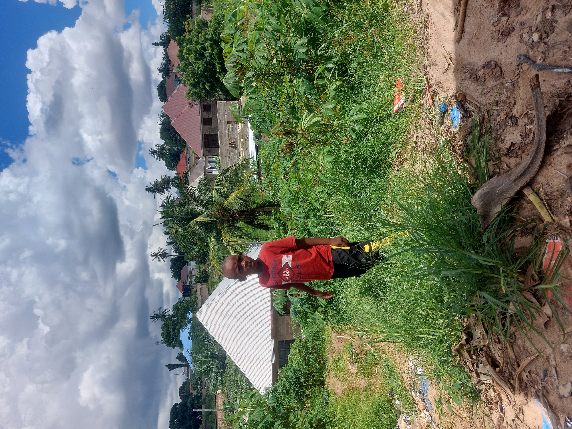 A young boy stands in a grassy area in Dar es-Salaam, Tanzania. He is wearing a red T-shirt and dark shorts. Behind him are houses and trees, and a cloudy sky is visible. The ground is uneven and has patches of dirt. There is some trash scattered nearby.