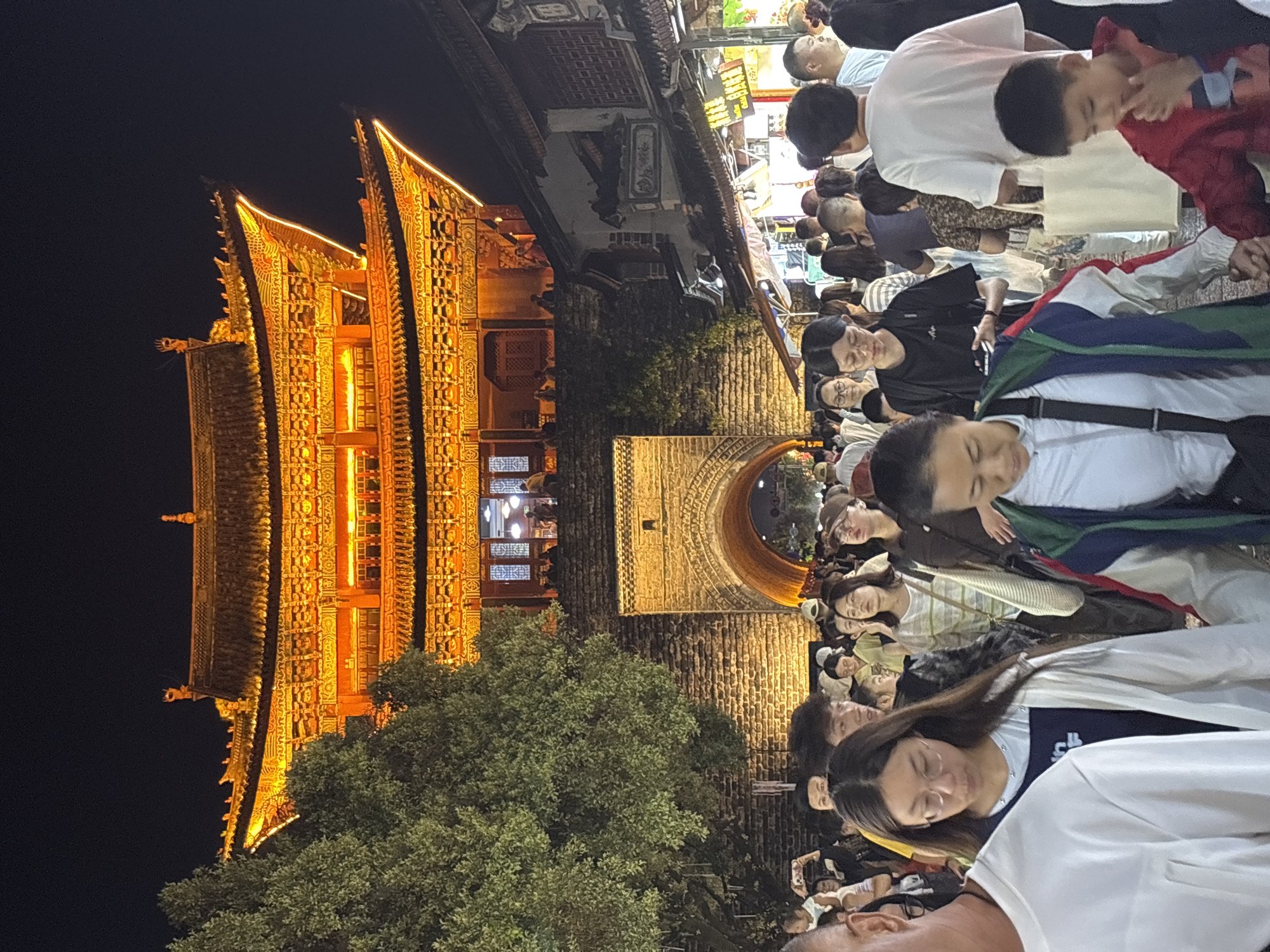 This image depicts a busy night scene in Dali City, China, likely at the Foreigner's Street (Yuan Yang Street). A large, illuminated traditional Chinese building, possibly a gate tower or temple, is visible in the background, adorned with intricate golden architecture and warm orange lighting. In the foreground and mid-ground, a dense crowd of people is walking, suggesting a lively marketplace or tourist area. The people are diverse in age and attire, with many looking at their phones or interacting with each other. The street appears to be paved, and there are stalls or shops lining the right side of the image, also lit up. A large tree with green foliage is situated to the left of the gate tower, providing a contrast to the urban setting. The overall atmosphere is one of a vibrant night market or cultural attraction, with the historical architecture serving as a prominent backdrop to the contemporary activity.