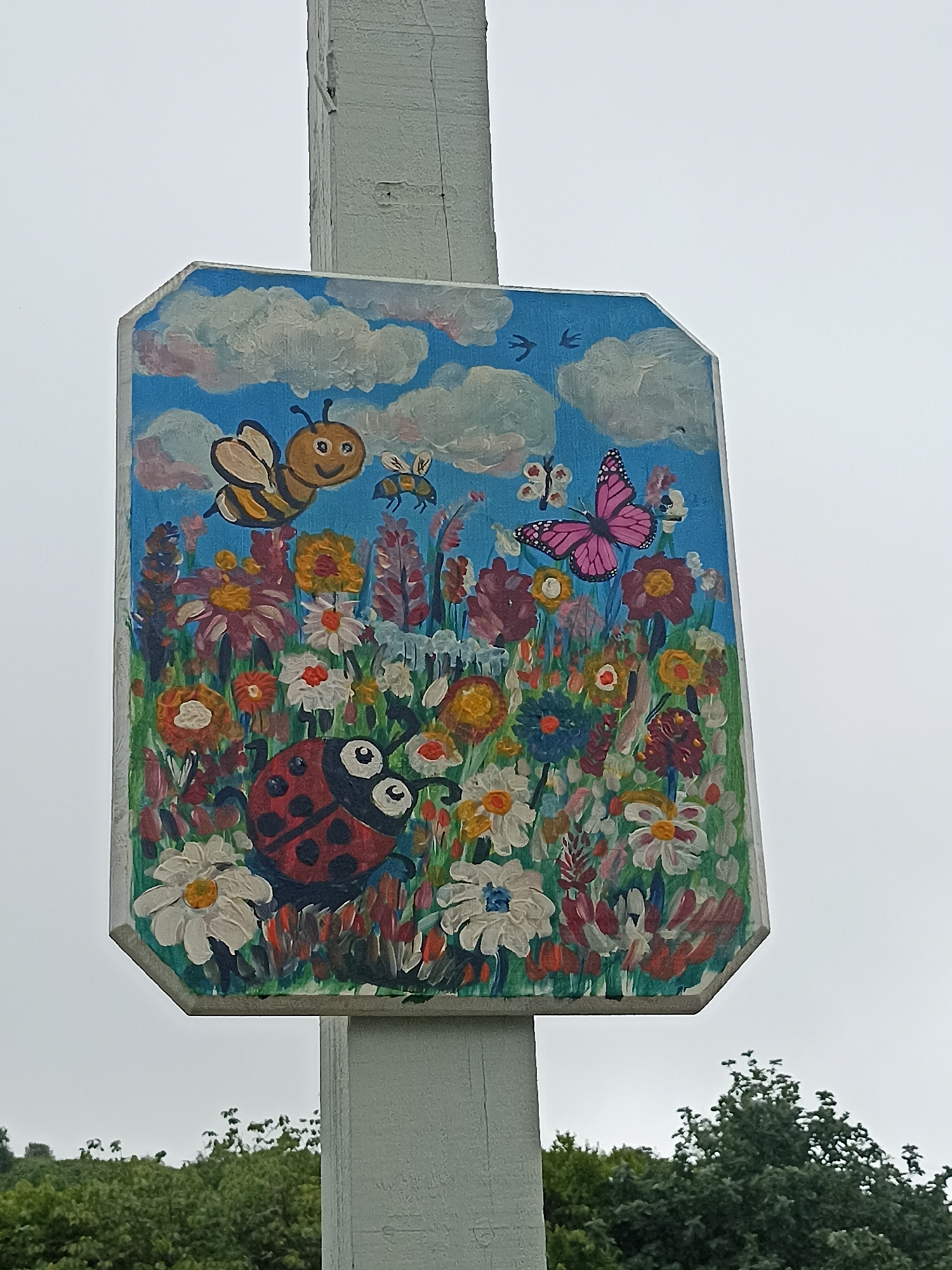 The image shows a close-up of a weathered, octagonal wooden sign painted with a colorful depiction of a garden scene. The sign is mounted on a light gray, rectangular post. The painting features a bright blue sky with white clouds and two small dark birds in flight. Below the sky, a vibrant field of flowers is depicted in various colors, including red, orange, yellow, pink, white, and blue. Prominent subjects in the painting include a large, cartoonish ladybug with black spots and wide eyes, a smiling bumblebee with striped yellow and black body and transparent wings, and a bright pink butterfly with black markings. Several smaller insects and flowers are also visible. The sign is slightly tilted and appears to be outdoors, with blurry green foliage visible at the bottom of the frame, suggesting it is in a garden or park setting. The background sky is overcast. The location is identified as an unknown location in Ireland.