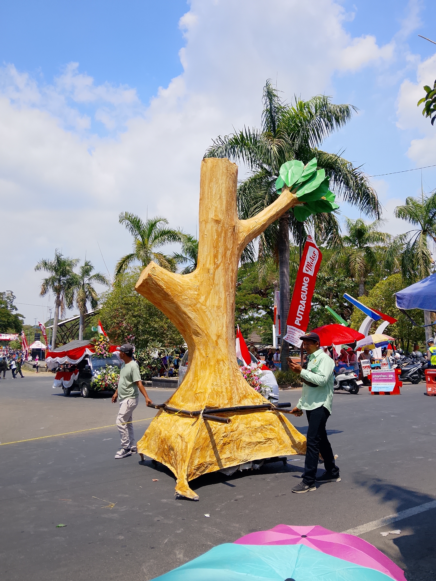 The image depicts a sunny outdoor scene, likely during a parade or festival in Balun, Indonesia, given the presence of palm trees and a truck decorated with flowers and a red and white canopy, resembling a float. The main subject is a large, stylized artificial tree trunk made of a golden-brown material, possibly paper mache or fabric stretched over a frame. Two men are interacting with this tree structure; one in a light green long-sleeved shirt and dark pants is holding what appears to be part of the tree's support system and looking at his phone, while another man in a light green shirt and light-colored pants stands near the base of the tree, also holding a part of the support. In the background, there are more palm trees, a red banner with white text that reads "PUTRAGUNG MALOR", and various other parade elements like flags and umbrellas, as well as spectators and parked motorcycles. The ground is paved asphalt. A pink and blue umbrella are visible in the foreground, partially obscuring the bottom of the frame.