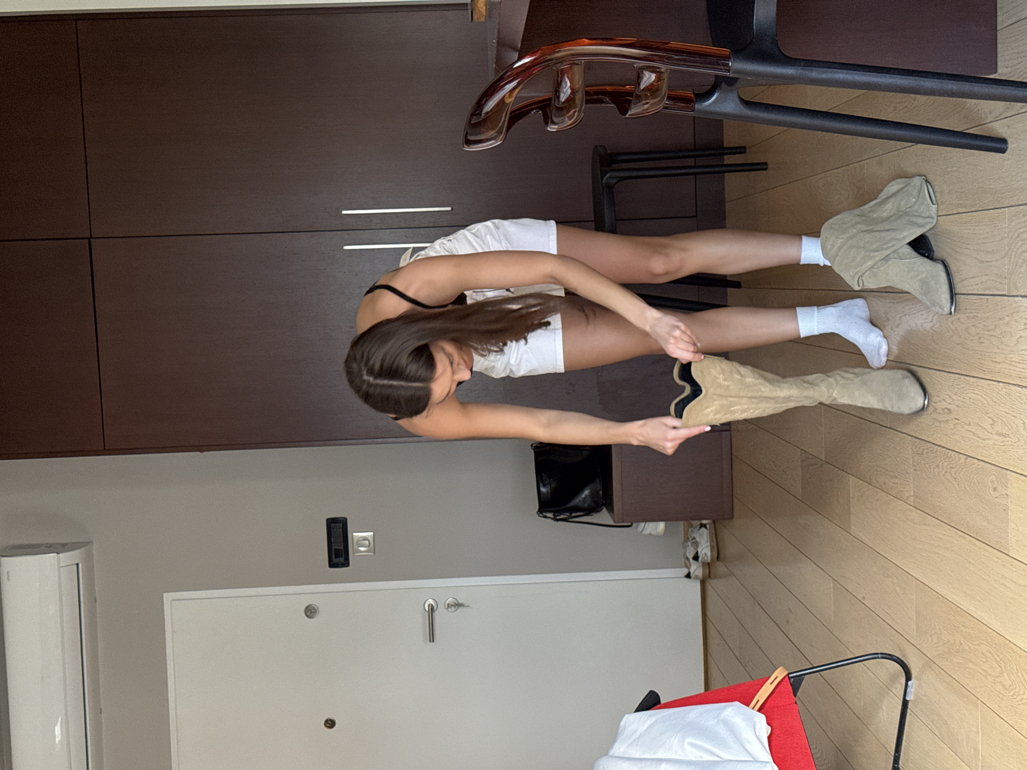 A woman wearing white shorts and white socks is putting on a tan knee-high boot in a room with a dark brown wardrobe against the wall. A chair with a wooden seat is to the right of her. In the background, there is a door, a light switch, and a radiator. The location is unknown, Unknown.