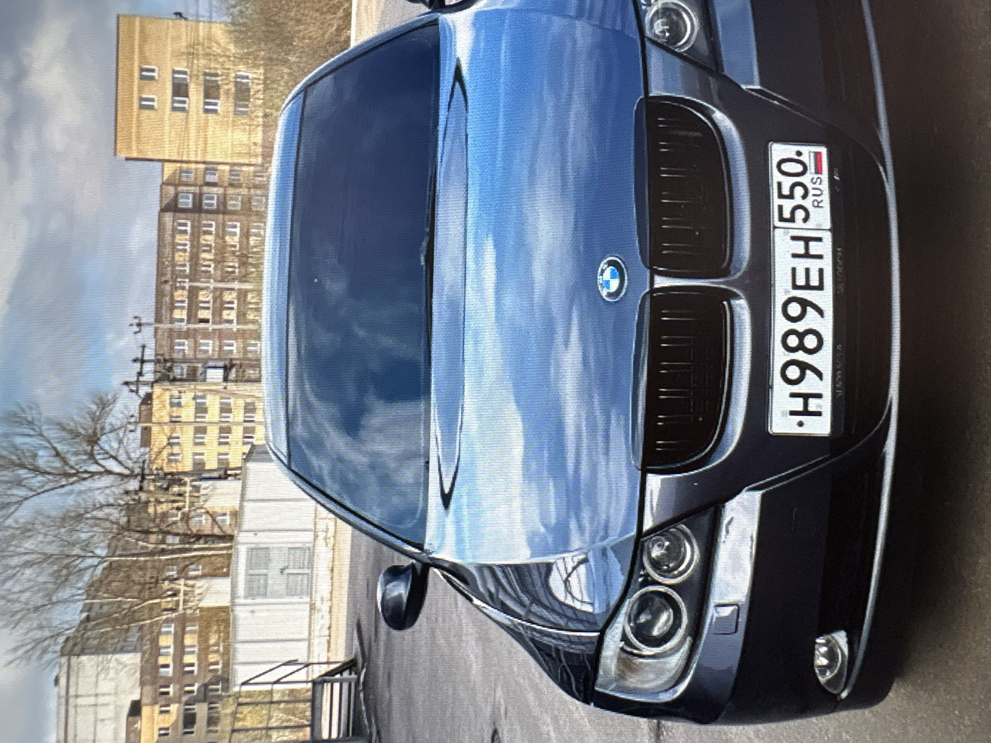 A dark blue BMW sedan is parked in front of apartment buildings in a city. The car's license plate reads "H989EH 550 RUS". The environment appears to be an urban setting with paved ground and a cloudy sky. The apartment buildings in the background are of various sizes and architectural styles, with some featuring yellow and beige facades. The image is taken from a slightly low angle, focusing on the front of the car. There are no people visible in the image. The scene appears to be a typical street view in a residential area.