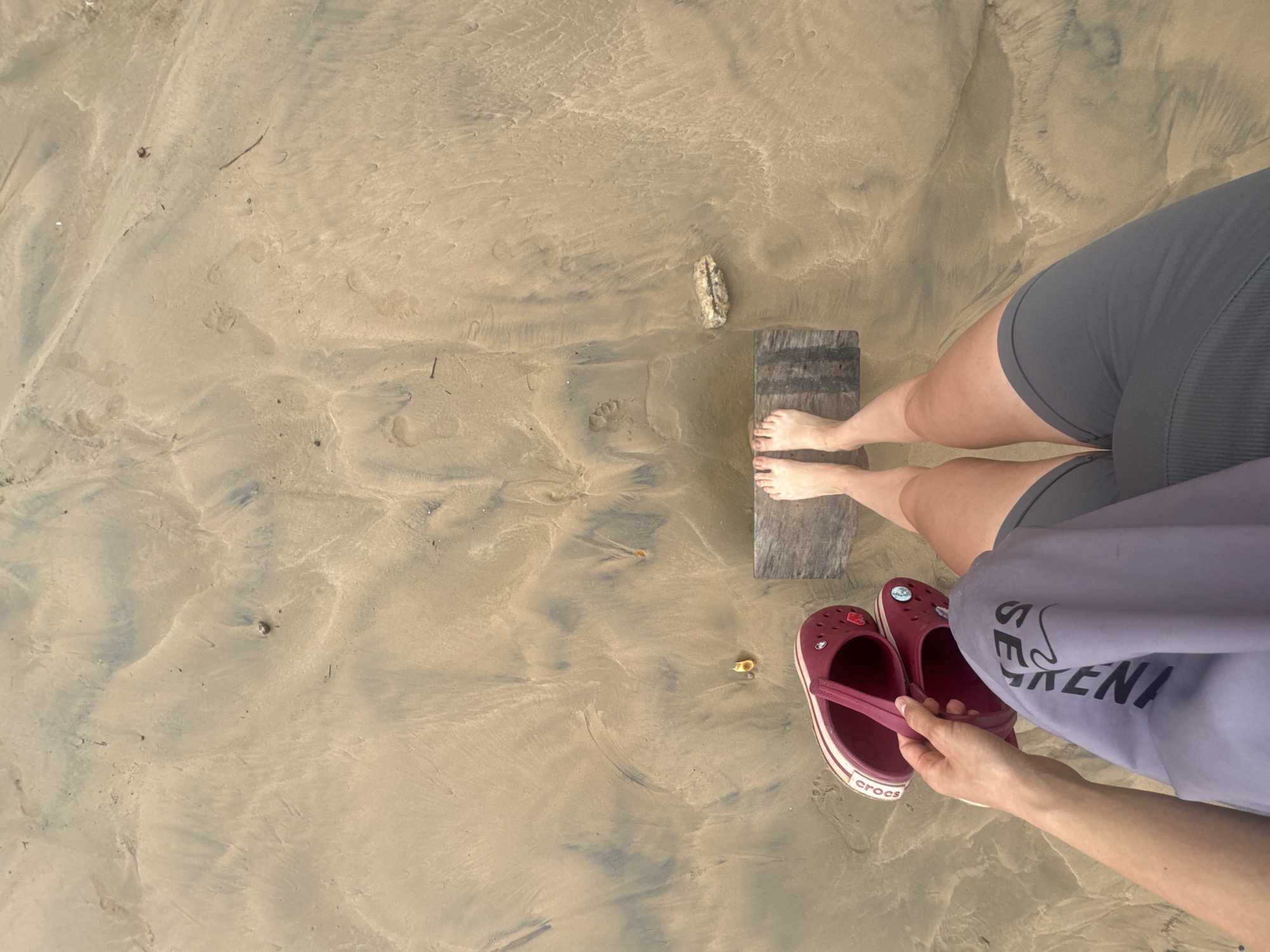 A woman stands on a sandy beach in Peleana, Sri Lanka. She is wearing gray athletic shorts and a light purple sweater. Her bare feet are on a piece of weathered wood. To her right are a pair of maroon Crocs sandals. The sand shows patterns of water movement and scattered debris. A small rock lies nearby. The view is an overhead perspective, likely taken from above.