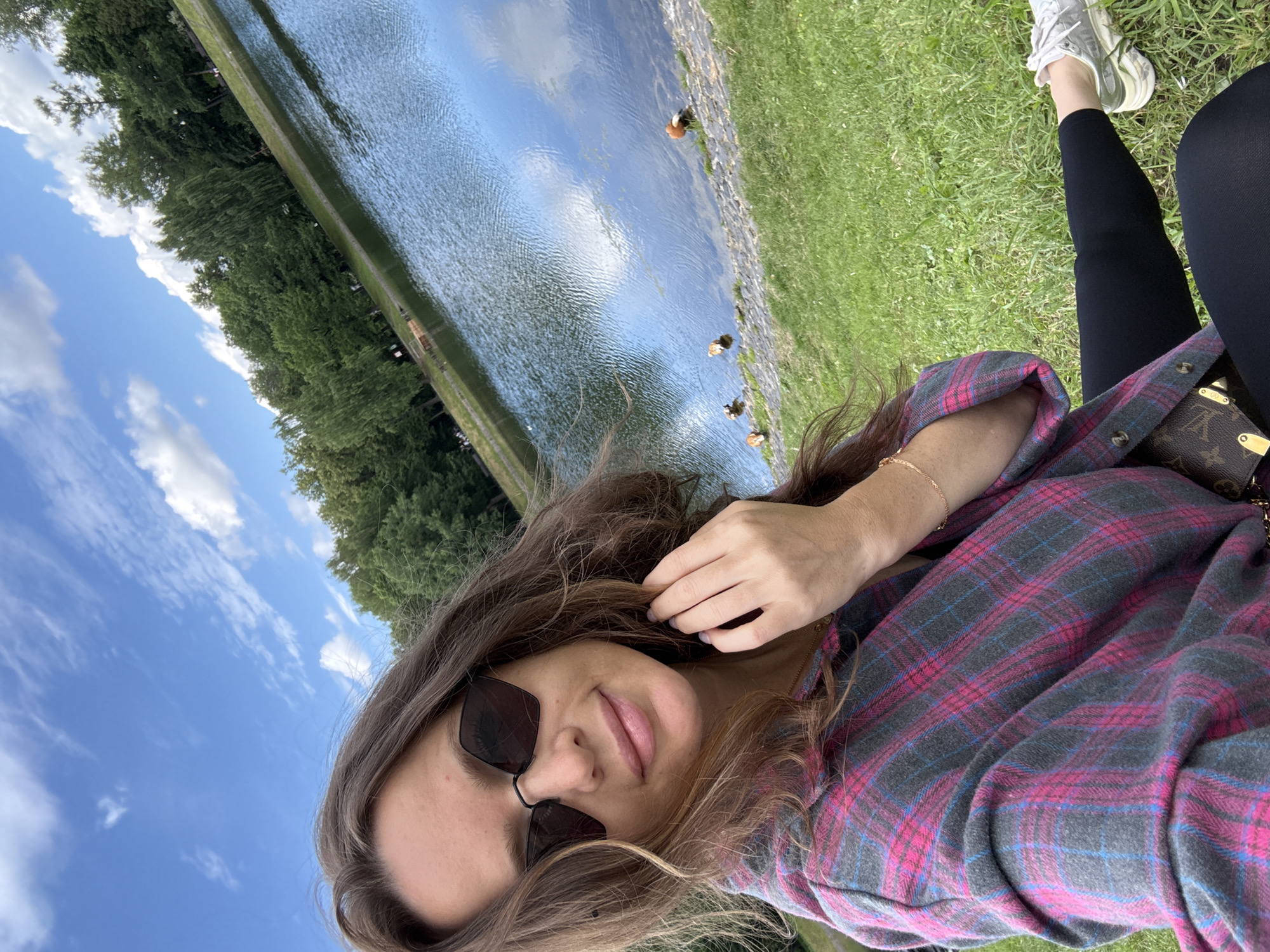 A young woman with long, wavy brown hair, wearing sunglasses, a pink and gray plaid shirt, black leggings, and white sneakers, is sitting on a grassy hill. She is looking at the camera and smiling, with her hand touching her hair. In the background, there is a calm lake surrounded by lush green trees and a bright blue sky with scattered white clouds. Several ducks are visible by the edge of the water. The scene appears to be a park or recreational area within the city of Moscow, Russia.
