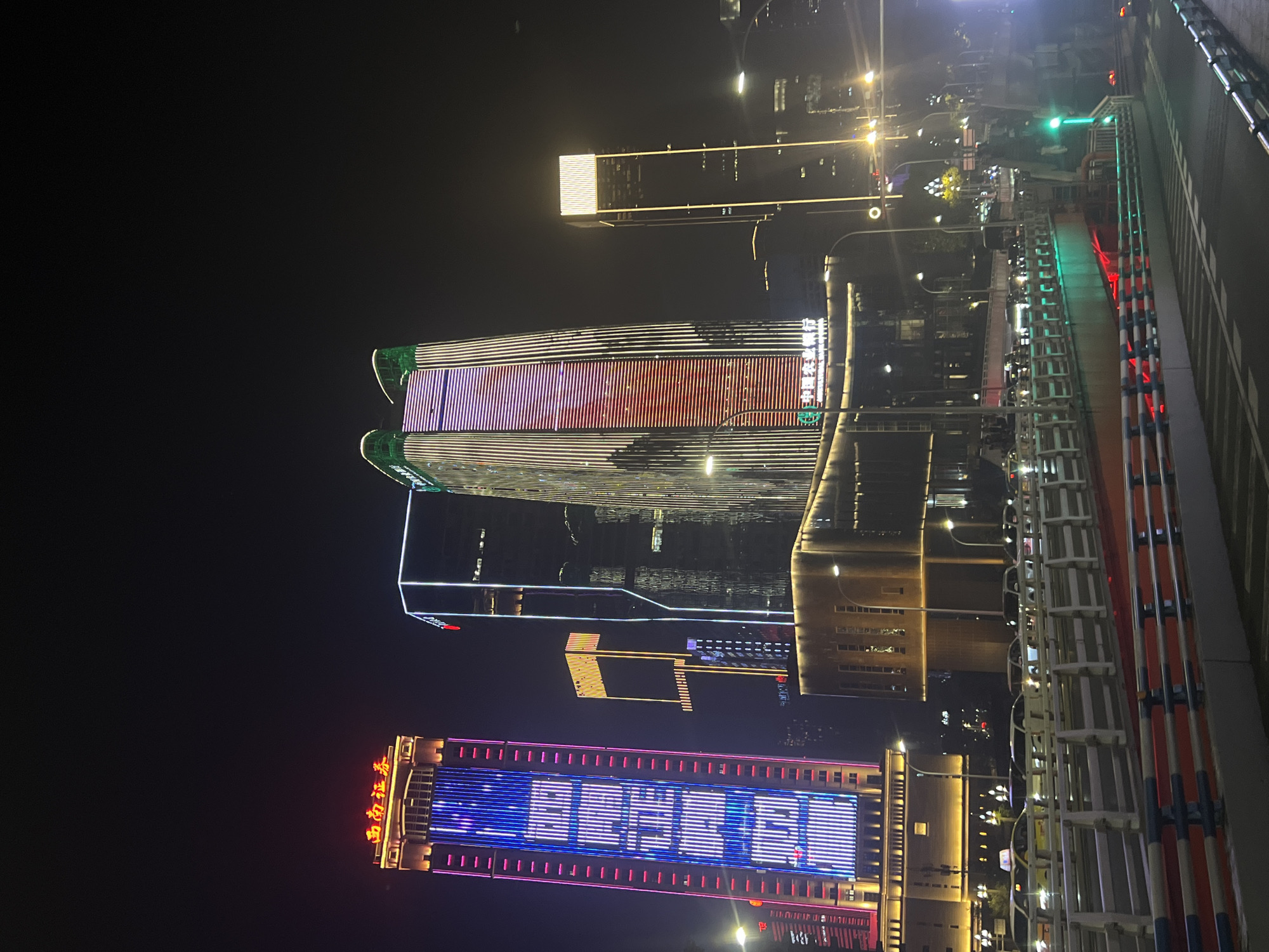 The image shows a nighttime cityscape with illuminated skyscrapers in Jiangbeicheng Subdistrict, China. The foreground features a multi-lane road or bridge with a reflective surface and metallic railings, indicating vehicle traffic. Several cars are visible on the road, some with headlights on. In the background, a cluster of modern buildings dominates the scene. One prominent skyscraper on the left displays vertical rows of purple and blue lights forming a pattern, with a red neon sign at the top reading "西南证券" (Southwest Securities). To its right, a taller building is illuminated with vertical white and green lights, and a section in the middle glows red. Another skyscraper to the far right is a slender, rectangular structure with a square illuminated top. The sky is dark, suggesting it is nighttime. The overall impression is a vibrant urban environment at night, characterized by modern architecture and extensive use of colorful lighting.