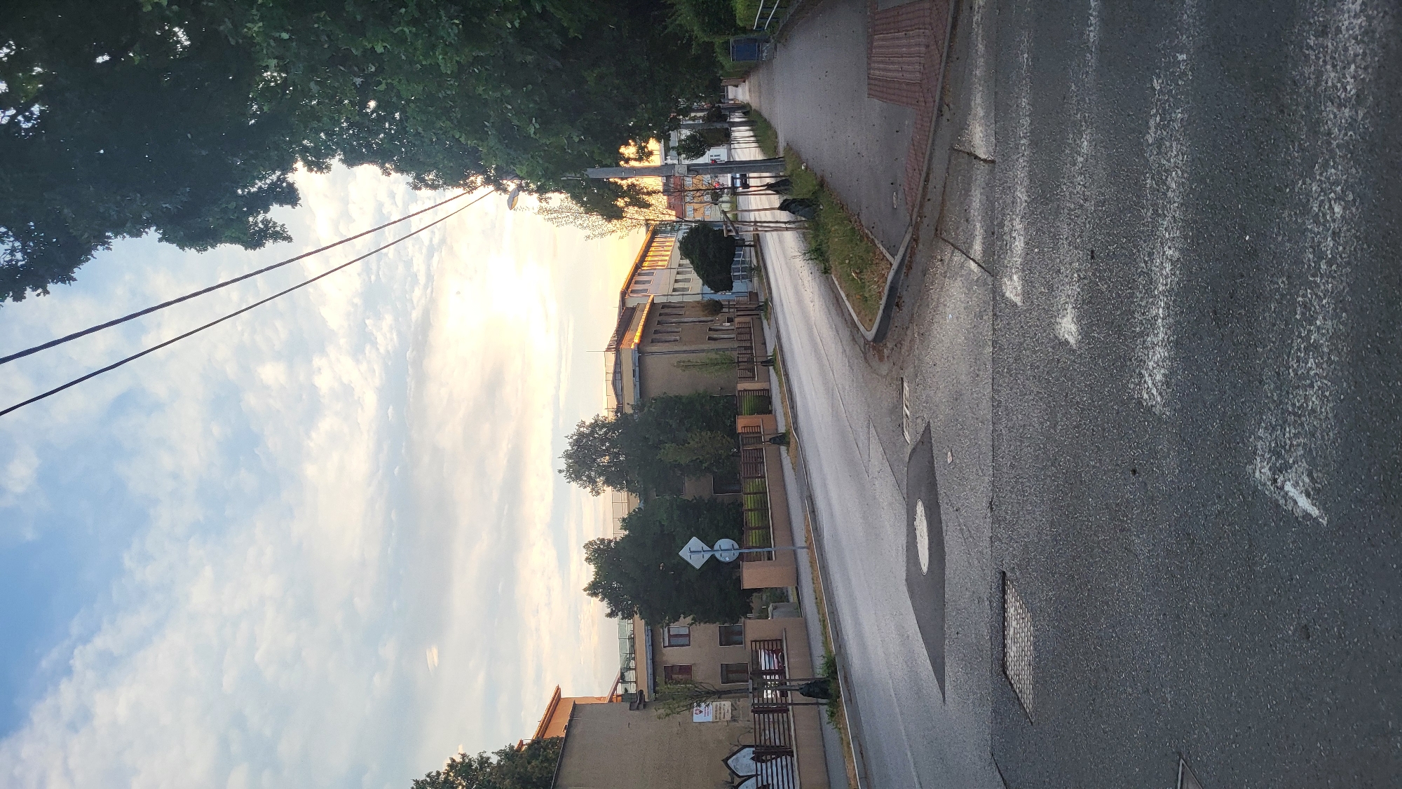 The image depicts a street scene in Prešov, Slovakia, at dusk. A paved road curves to the right, with a sidewalk on the right side. Several buildings, appearing to be educational or institutional, line the left side of the road. Trees are present on both sides of the street and in front of the buildings. Overhead power lines are visible against a sky with scattered clouds and the setting sun. The road shows some wear and tear, with markings and manhole covers visible. There are no people or vehicles immediately apparent in the scene.