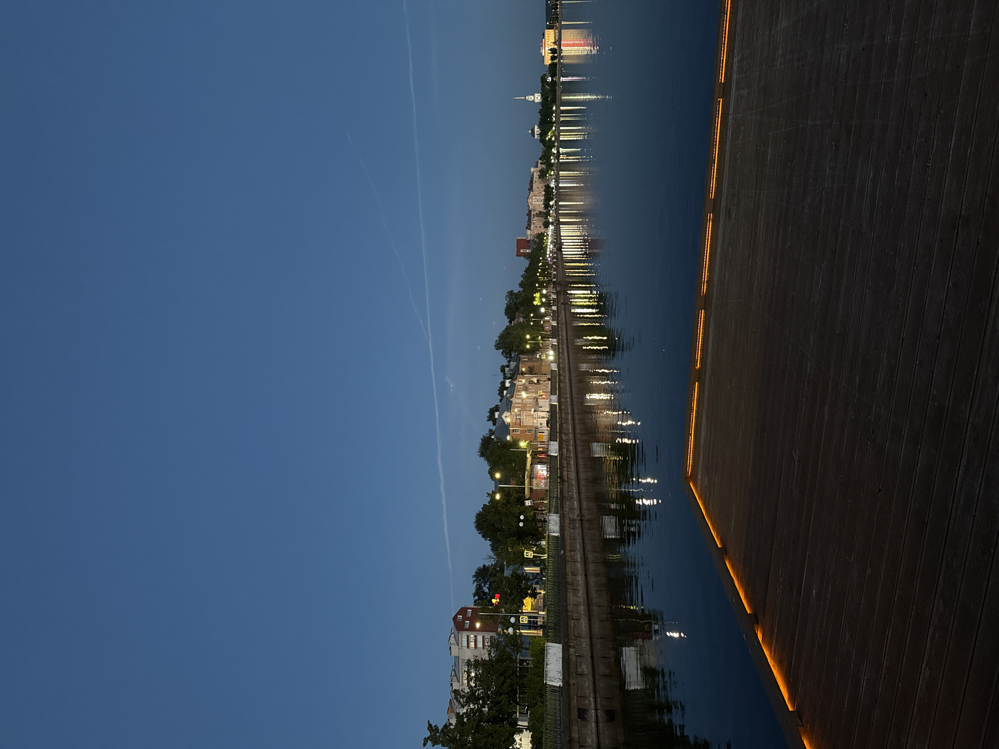 This image presents a serene nighttime view of a waterfront in Votkinsk, Russia. The scene is illuminated by artificial lights, reflecting beautifully on the calm surface of a body of water, likely a river or lake.
In the background, across the water, the skyline of a town or city is visible. Various buildings, including residential structures and what appears to be a white church-like building with a distinctive dome and spire, line the distant bank. A promenade along the waterfront is lit by numerous streetlights, creating vibrant, elongated reflections that stretch across the dark water. Patches of trees punctuate the urban landscape.
The sky above is a deep, clear blue, indicative of twilight or early night. A faint, thin white contrail from an aircraft is discernable high in the sky.
In the foreground, on the right side, a modern wooden boardwalk or pier extends into the scene. It is constructed from dark planks and features embedded orange LED strip lighting along its edge, casting a warm, inviting glow that contrasts with the cooler tones of the water and sky. These lights also create gentle, shimmering reflections on the water surface close to the pier.
There are no people or specific activities taking place, contributing to the tranquil and contemplative mood of the scene. Distant signs on the opposite bank are too far and indistinct to be legible. The overall impression is one of peaceful evening beauty, showcasing the interplay of urban architecture, light, and water.