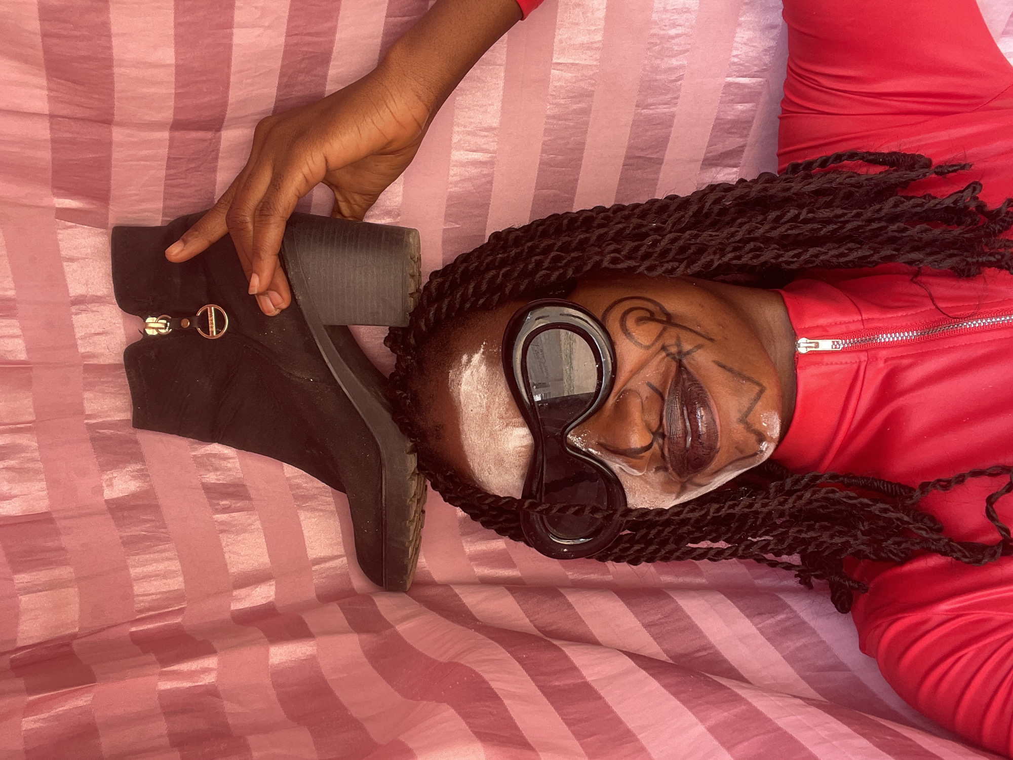 A person in a red garment lies on a pink and white striped fabric. The person wears round sunglasses and face paint. They have long dark braids. A hand holds a black ankle boot with a "D" ornament on the zipper. The location is unknown, and there is no information to suggest that this image was taken in Akwanga, Nigeria.