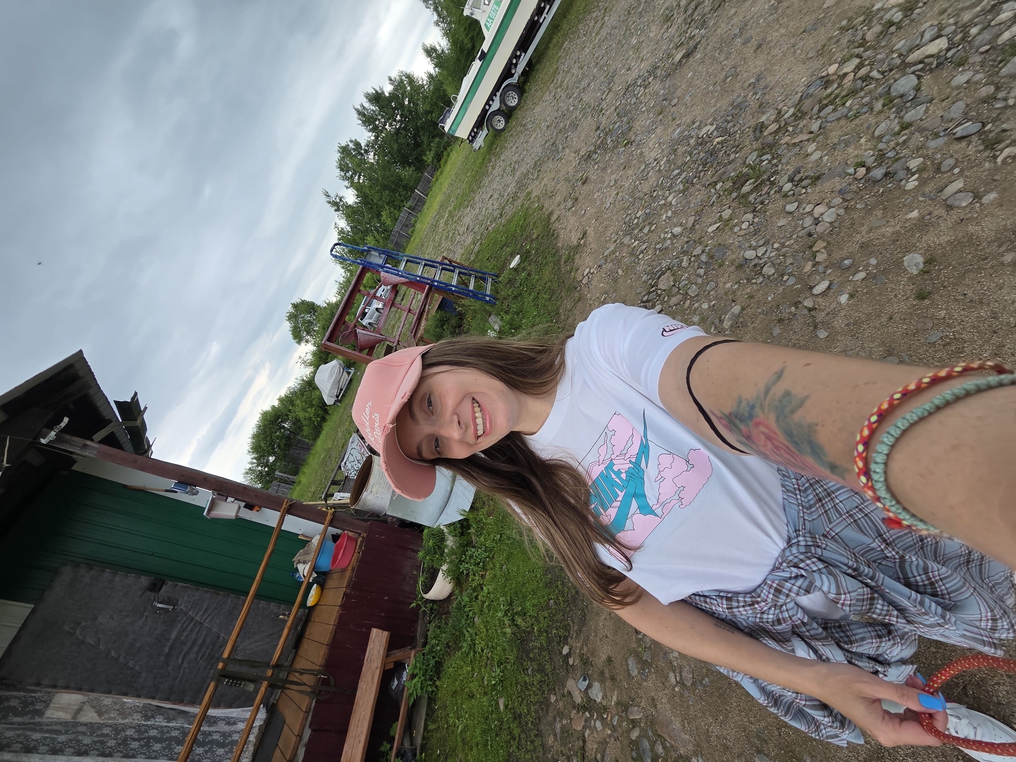 In Talakan, Russia, a woman with brown hair, wearing a pink baseball cap, a white t-shirt with a graphic print, and a plaid shirt tied around her waist, stands in a gravelly yard. She is smiling and holding a red rope. Her left arm displays a tattoo, and she wears bracelets on her wrist. Behind her, a small wooden house with green siding and a porch is visible, along with various objects like buckets and a wooden bench. Further in the background, a boat on a trailer, a metal structure with a ladder, and trees under a cloudy sky complete the scene.