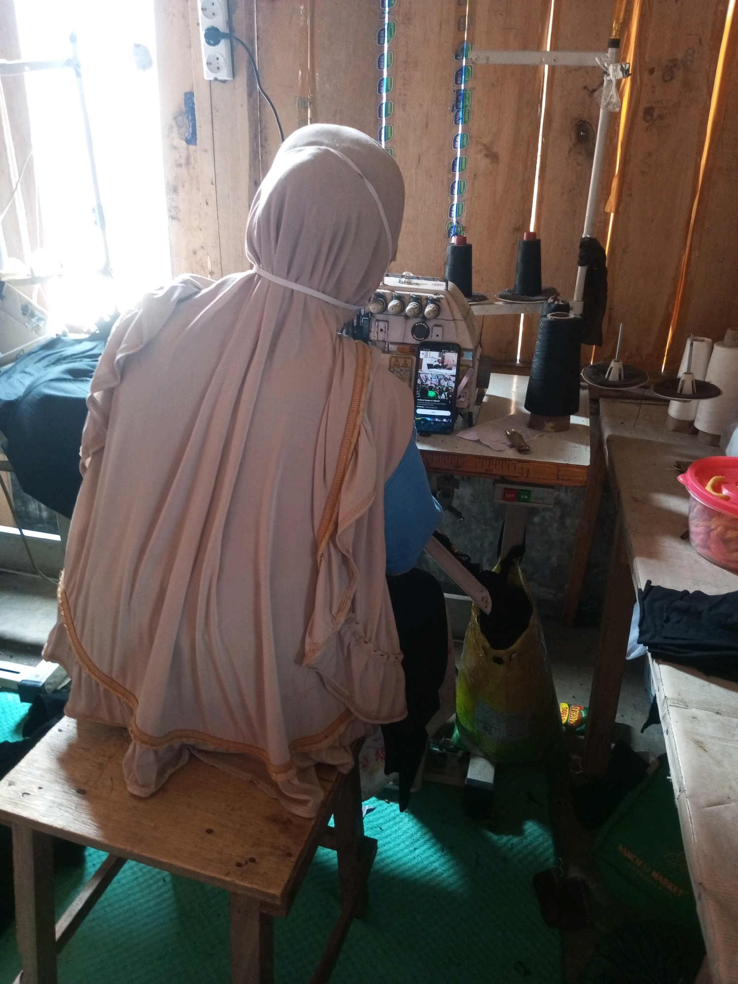 In the city of Kalibeber, Indonesia, a person wearing a light-colored headscarf and a light-colored garment with ruffled edges is seated on a wooden stool in front of a sewing machine. The person's face is not visible. The sewing machine is positioned on a wooden table, and a smartphone is propped up on the machine, displaying a video. Several spools of black thread are visible on the sewing machine. Behind the sewing machine, a wooden wall is present. To the left of the sewing machine, a window with bright light streaming through it is visible. A dark blue fabric is draped over something to the left. On the table to the right of the sewing machine, there are spools of white thread and a red plastic container filled with items. A large yellow bag and a green bag are on the floor. The floor is covered with a green mat. The overall scene depicts a sewing workspace.