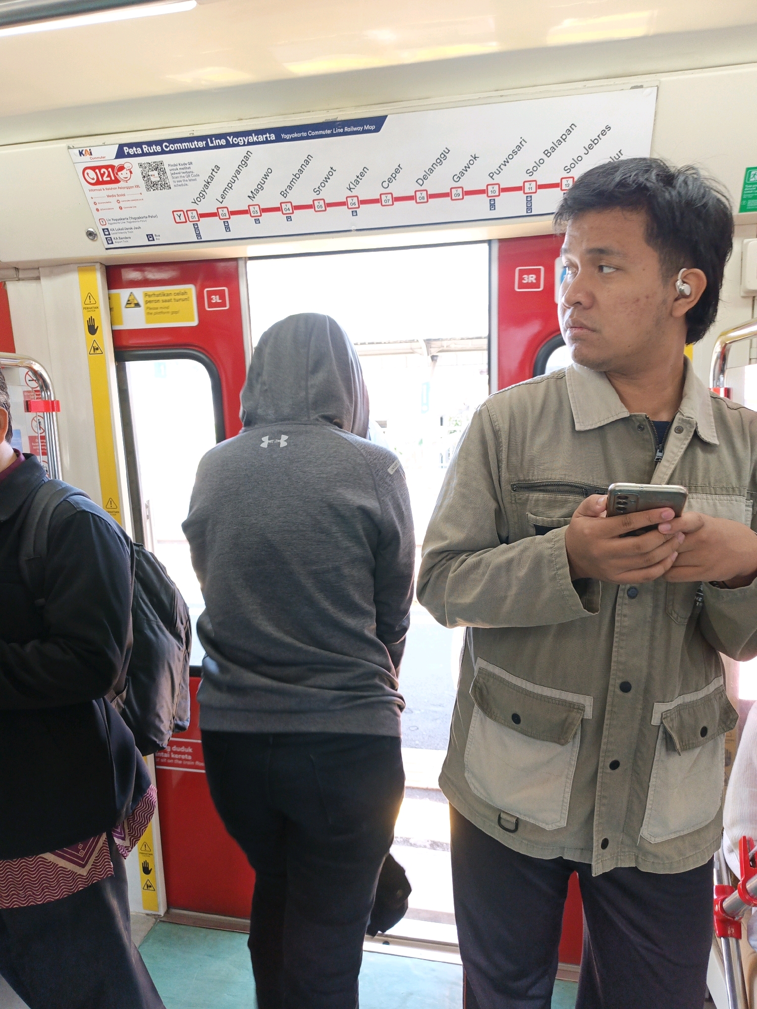 This is a medium shot, eye-level, indoor photograph taken inside a train carriage, likely a commuter train given the map displayed. The lighting appears to be bright, possibly from the sun outside the train.
In the foreground on the right, a young man with short, dark hair and a light brown jacket is looking to the right, away from the camera. He is holding a smartphone in his right hand and appears to be using it. He has an earbud in his right ear. The jacket has two chest pockets with flaps and two lower pockets, one on each side. He is wearing dark pants.
To the left of the young man, a person wearing a grey hooded sweatshirt with the Under Armour logo is facing away from the camera, towards the train door. They are wearing dark pants.
Further to the left, a person wearing a dark jacket over a garment with a patterned hem is partially visible.
Above the passengers, a train route map is displayed. The map is titled "Peta Rute Commuter Line Yogyakarta" and shows various stations, including Yogyakarta, Lempuyangan, Maguwo, Prambanan, Srowot, Klaten, Ceper, Delanggu, Gawok, Purwosari, Solo Balapan, and Solo Jebres. There are also QR codes and other information displayed on the map.
The train carriage interior is visible with red doors and yellow safety strips on the sides. The floor is a light green color. The overall scene depicts a moment of travel on a commuter train in Indonesia, with the specific location potentially being the Yogyakarta Commuter Line.