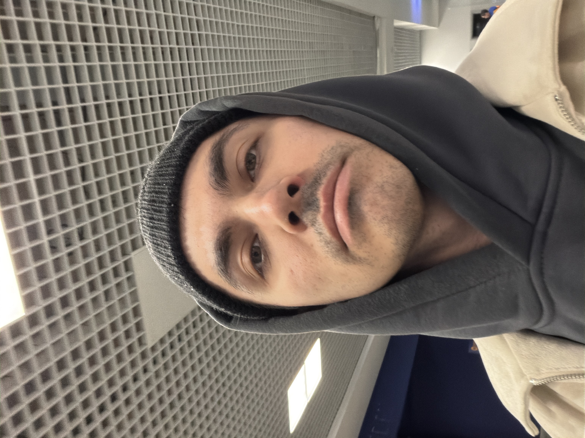A close-up photograph shows a person wearing a gray hooded sweatshirt and a gray knit hat under the hood. A beige jacket is partially visible over the sweatshirt. The person's face is turned slightly, with eyes and nose visible. The background consists of a grid-like ceiling. The location of the photograph is not discernable in the image. The city of Izhevsk, Russia, is not confirmed by the image.