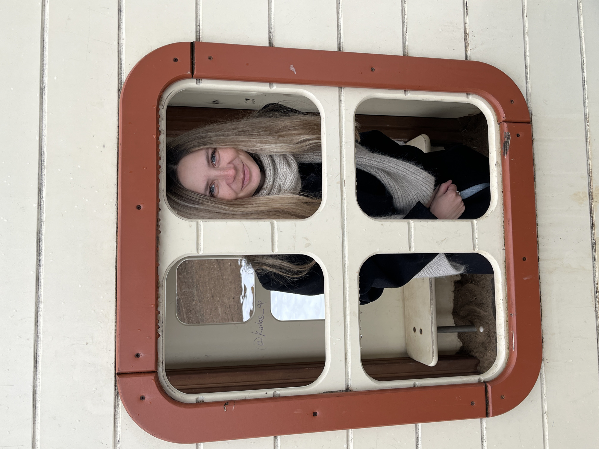 A woman with long blonde hair is peeking through a window. The window frame is white with a brown border. The window has four panes, and the woman's face fills the upper left pane. She is wearing a black coat and a gray scarf. The scene appears to be set in Chaikovsky, Russia.