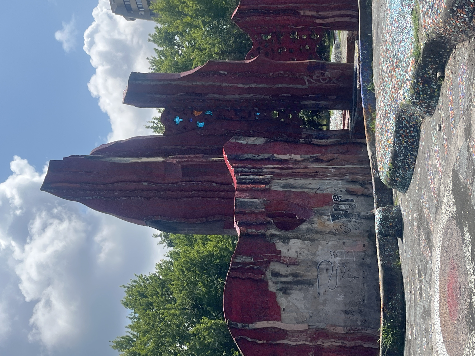 The image shows a public art installation in Sochi, Russia. The installation is made of red, textured material that appears to be concrete or mosaic tile, with some sections showing wear and graffiti. It features abstract, vertical forms resembling rocky outcrops or organic structures. There are some colorful mosaic elements and what appear to be blue and orange glass inserts on one of the central structures. The foreground includes a circular area with red and white mosaic patterns and curved, low walls also covered in colorful mosaics. Green trees form a backdrop to the installation, and a modern building is visible in the upper right corner. The sky is partly cloudy, with bright sunlight. There are no people visible in the image.