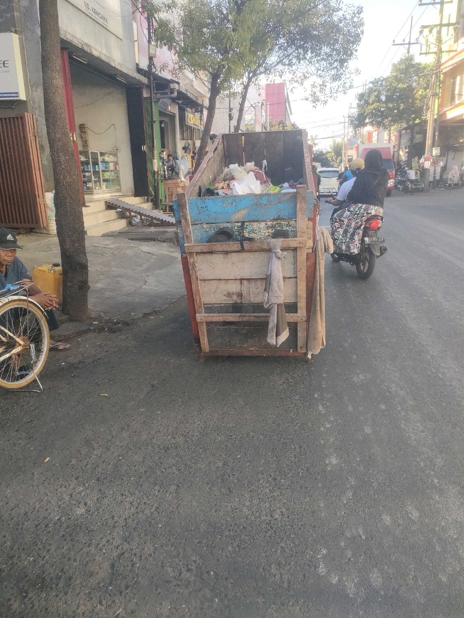 In Surabaya, Indonesia, a man sits beside a bicycle and a yellow container on a paved street. In the background, a dumpster filled with trash is visible. A person rides a motorcycle past the dumpster. Buildings line the street, with a tree in the foreground. The sky is bright and sunny.