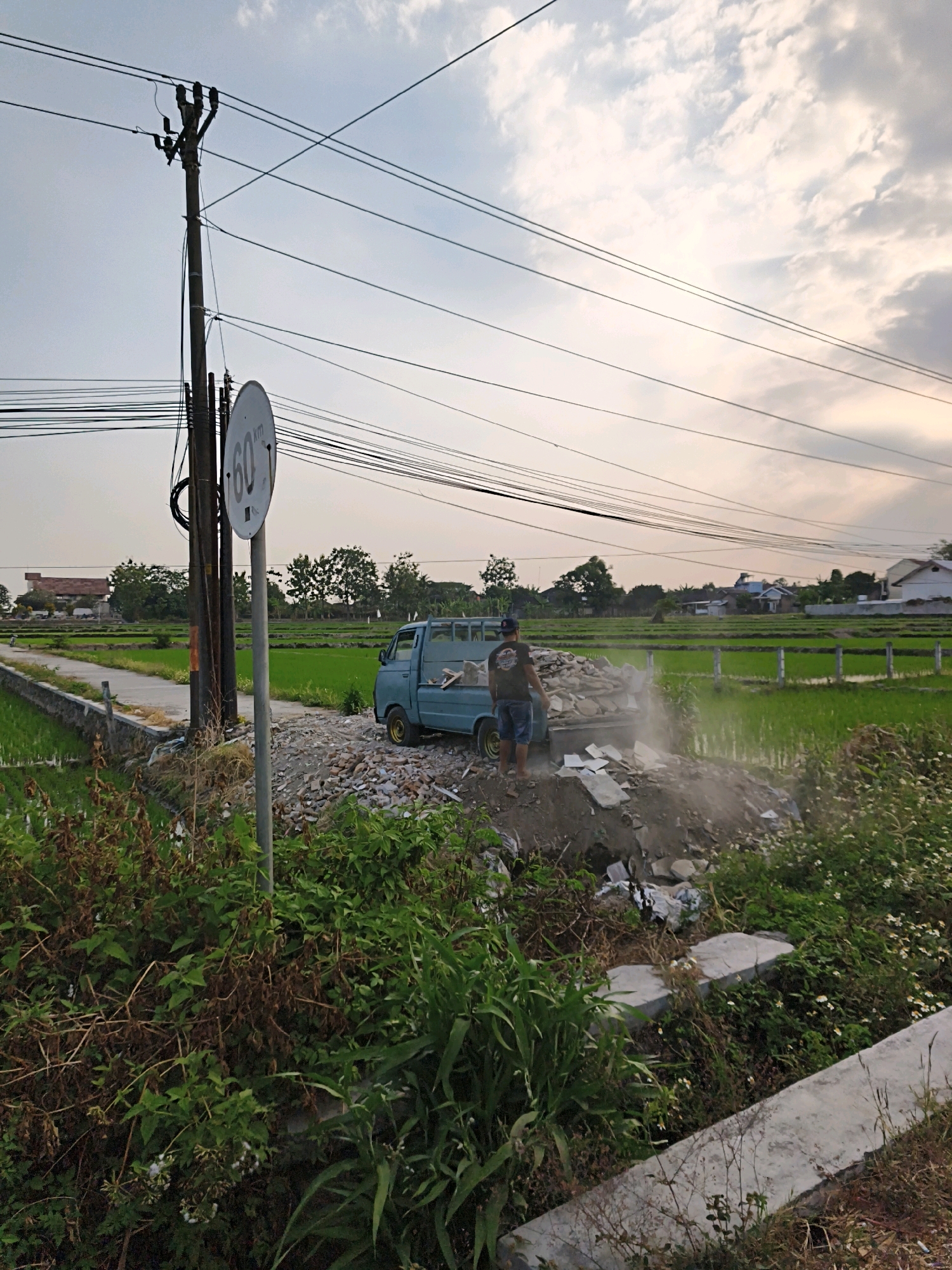 An overcast, late afternoon scene in Cangkringan, Indonesia, shows a light blue pickup truck parked on a dirt and rubble-strewn area adjacent to a paved road and a vibrant green rice paddy. A person, wearing a dark t-shirt, shorts, and a baseball cap, stands on the edge of the truck's bed, appearing to be in the process of unloading or disposing of debris, which consists of broken concrete and other construction-like materials. Dust is visibly rising from the pile.
Dominating the left side of the image is a wooden utility pole with multiple electrical wires crisscrossing the sky. A circular speed limit sign, indicating "60 KM," is mounted on a metal pole in front of the utility pole. The background features a row of trees and houses, with the rice paddy extending into the distance. Vegetation, including tall grasses and leafy plants, lines the foreground and the edge of the road. The overall impression is of a rural or semi-rural setting where construction debris is being managed.