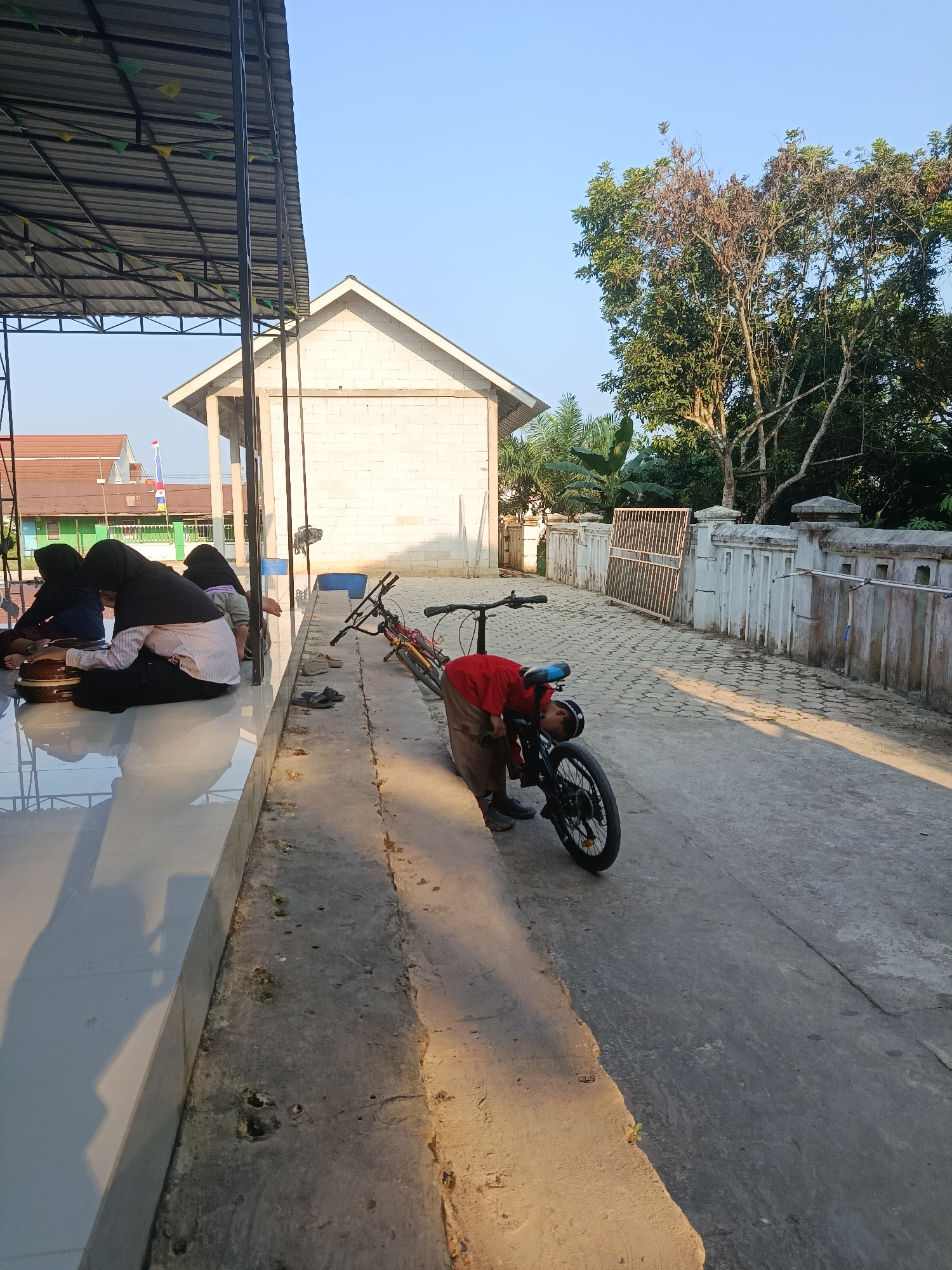 In Swarga Bara, Indonesia, this image captures a scene outdoors under a clear blue sky, likely during daylight hours. In the foreground, to the left, three individuals wearing hijabs are seated on a light-colored tiled surface beneath a metal-roofed structure supported by dark vertical poles. One individual is interacting with circular objects. To the right of the seated individuals, a young person in a red shirt and brown pants is bending over a bicycle that is lying on its side on a concrete pathway. The bicycle is black with blue accents on the seat and has visible spokes and a rear wheel. Two pairs of sandals are also present on the concrete near the bicycle. The background features a white brick building with a gabled roof, followed by trees and a white stone fence with decorative elements. A metal gate is visible further down the pathway. The environment appears to be a public area, possibly near a school or community center, given the building and the presence of children.