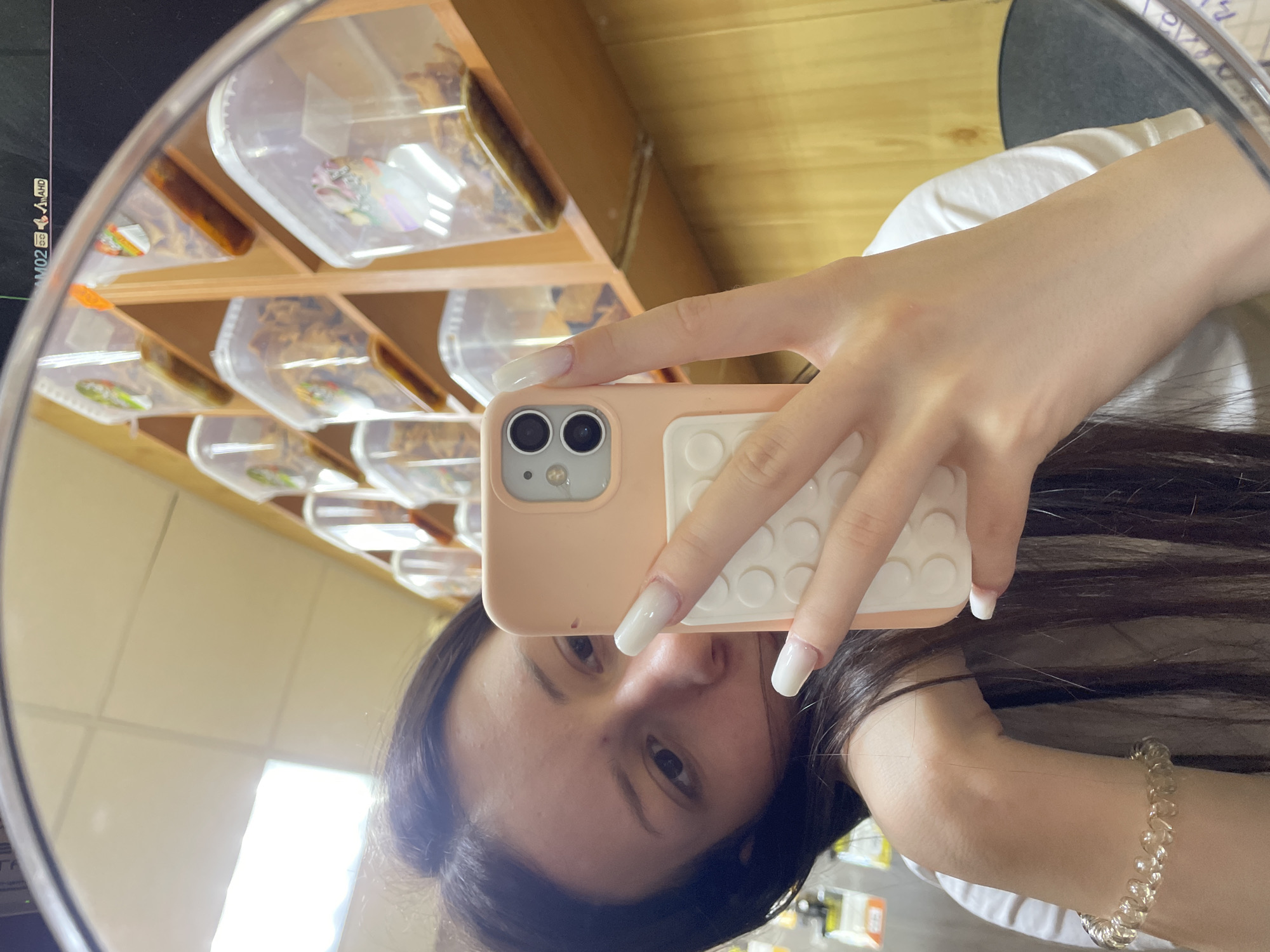 A sideways mirror selfie captures a young person holding a light peach-colored smartphone with a white, textured grip. The individual's face is partially obscured by the phone, revealing one eye and a portion of their forehead and long, dark hair. Their right hand, adorned with long, light-colored nails, holds the phone. A clear, spiral-shaped bracelet is visible on their right wrist. They are wearing a white t-shirt.
The reflection in the mirror shows an indoor setting, likely a shop or pantry. In the background are wooden shelves stacked with numerous clear plastic containers. These containers appear to hold various dried goods, possibly nuts, seeds, or candies, with some featuring small colorful labels or images. A bright light source is reflected in the upper left of the mirror, suggesting it's daytime. Faint text and symbols, including "2M02", "AHD", and "HD", are visible on a screen or surface reflected in the far left of the mirror.
