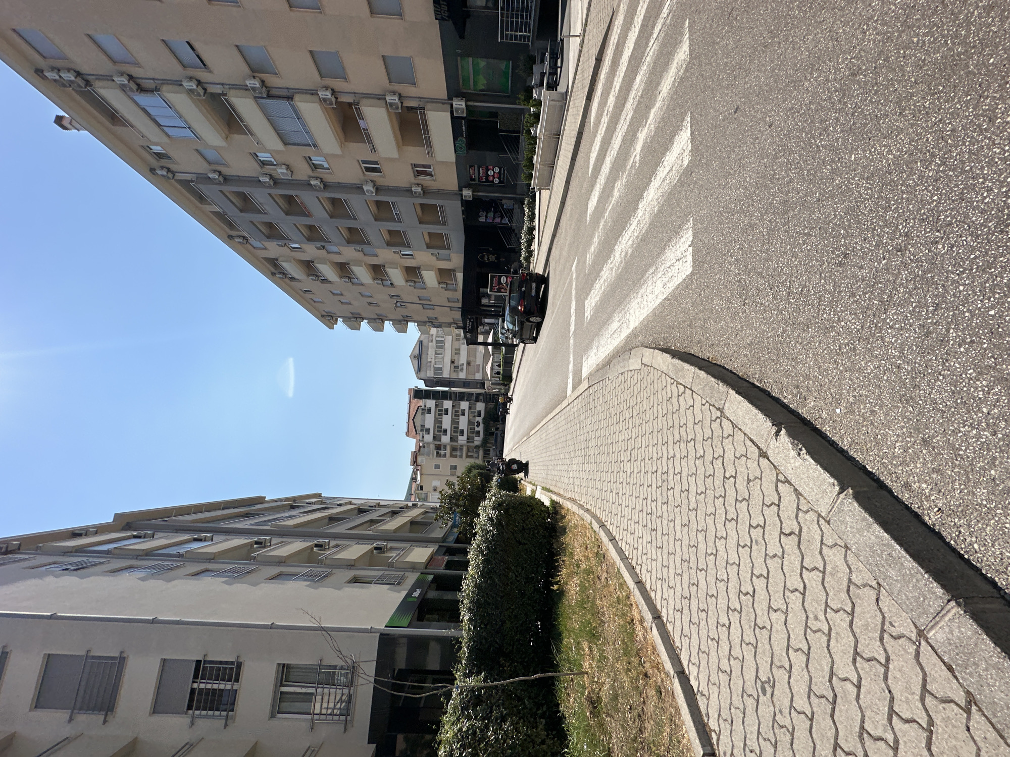 The image depicts a street scene in Budva, Montenegro, under a clear blue sky, suggesting a sunny day. The perspective is from a low angle, looking up and along the street, which is lined with modern apartment buildings on both sides. The buildings are a mix of light beige and grey, with numerous windows and air conditioning units visible. Some buildings have balconies, and others appear to have commercial spaces on the ground floor with large windows and signage.
A prominent feature of the scene is a pedestrian crossing painted in white stripes on the asphalt road. To the right of the crossing, there's a sidewalk paved with interlocking concrete blocks. Adjacent to the sidewalk, a neatly trimmed hedge separates the pedestrian area from the road.
In the distance, on the road, a black car is stopped, possibly at the pedestrian crossing. Further down the street, more buildings and a glimpse of the sky are visible. The overall impression is of an urban environment in daylight, possibly in the late morning or early afternoon due to the bright sunlight and clear sky. No specific activities are clearly taking place beyond the presence of the car and the implied use of the pedestrian area.
One building on the left has a green sign with white text that appears to say "T-COM". Another sign on the right building shows "LOTTO" and other text that is partially obscured. The scene conveys a sense of everyday urban life in a well-maintained area.