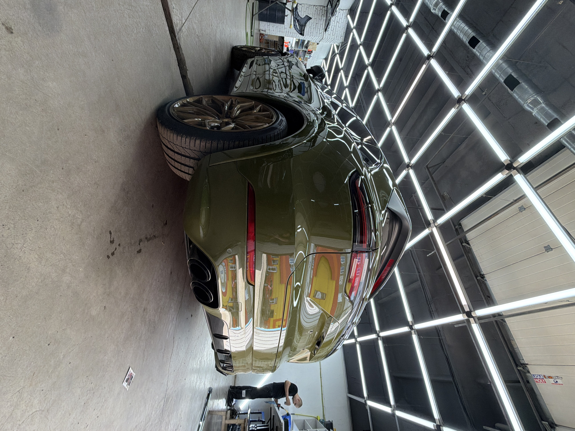 The image shows a dark green Porsche Cayenne parked inside a well-lit garage, likely a car detailing or customization shop. The garage features a grid of fluorescent lights on the ceiling and exposed ventilation ducts. The car's rear is prominently displayed, showcasing its sleek design, quad exhaust pipes, and large gold-colored wheels. A person wearing dark clothing and a black cap is standing to the right of the car, appearing to be working on something at a table. The floor is concrete, and there are some tools and equipment visible in the background, including shelves and what looks like a car detailing station. There are reflections on the car's body, indicating a glossy finish. The image does not contain any specific landmarks or clues that would allow for a precise location within Krasnoyarsk, Russia.
