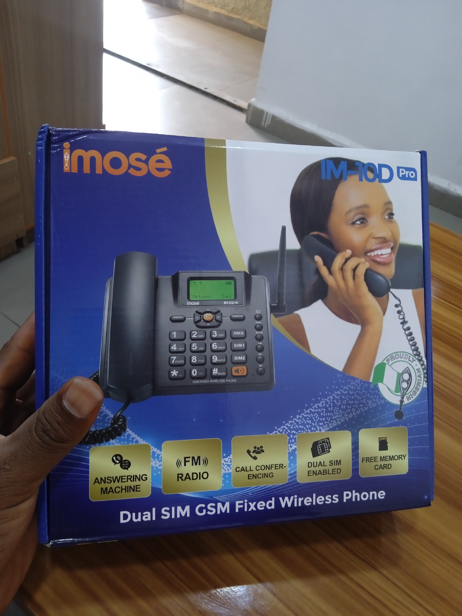 Here is a description of the image:
A person is holding a blue box that contains an "imose IM-10D Pro" Dual SIM GSM Fixed Wireless Phone. The box shows the phone with an antenna, along with a woman talking on the phone. The box also has the features of the phone described on it: answering machine, FM radio, call conferencing, dual SIM enabled, and a free memory card. The box shows a symbol which reads: "PROUDLY NIGERIAN". The background is out of focus, but there is a desk.