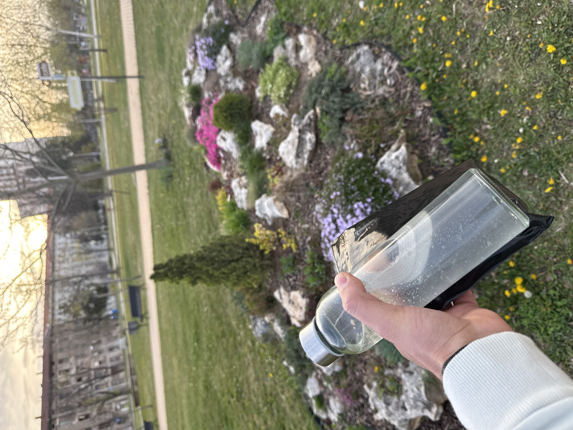 A slightly tilted, first-person perspective image captures a hand holding a clear bottle against an outdoor background in Sofia, Bulgaria.
**Subjects:** The primary subject in the foreground is a person's right hand, clad in a white long-sleeved top with ribbed cuffs, holding a transparent glass or plastic bottle. The bottle contains a clear liquid with small bubbles and is partially enclosed in a black sleeve. The background features a vibrant rock garden, green grass, trees, and distant urban structures.
**Scene and Setting:** The setting is a park or garden within an urban environment. In the mid-ground, a rock garden displays various low-lying plants and small flowers in shades of pink, purple, and yellow, interspersed with light-colored rocks. To its left, a small coniferous shrub is visible. Beyond the garden, a well-maintained grassy area extends, dotted with yellow dandelions. A light-colored paved path runs horizontally across the scene. In the distance, bare deciduous trees stand alongside streetlights, and multi-story buildings are faintly visible, indicating an urban landscape.
**Activity:** The activity is simply a person holding a bottle outdoors, likely during a walk or break in the park.
**Notable Details:** The time of day appears to be late afternoon or early evening (golden hour), given the warm, soft light illuminating the scene, particularly the sky and distant buildings. The weather is clear and sunny. There are no clear visible emotions on the unseen person.
**Visible Text:** No discernible text is visible on any object in the image.
