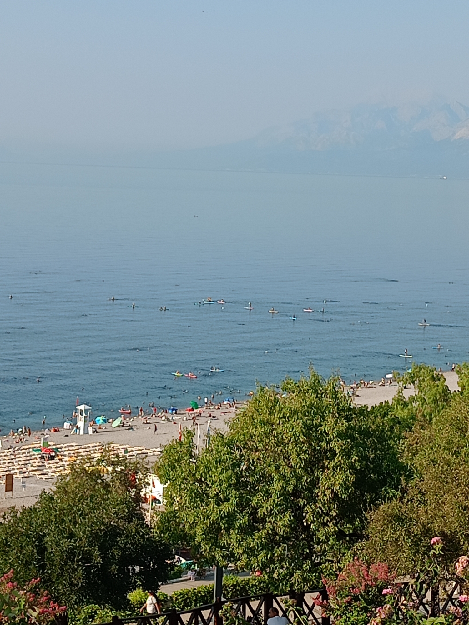 This is an aerial, full-shot photograph taken on a hazy, sunny day, overlooking a busy beach and the calm, blue sea. In the foreground, dense green trees and flowering bushes partially obscure the view of a dark wooden fence and a paved walkway with a few individuals walking. Further back, the sandy beach is crowded with sunbathers, lounge chairs, and umbrellas. A white lifeguard tower stands prominently on the beach, with a red-topped boat nearby. The sea is dotted with numerous people enjoying various water activities, including many on paddleboards and a few in small boats. In the distance, hazy blue mountains rise from the horizon. The scene captures a lively summer day at a coastal location, likely within Antalya, Turkey, given the landscape.
