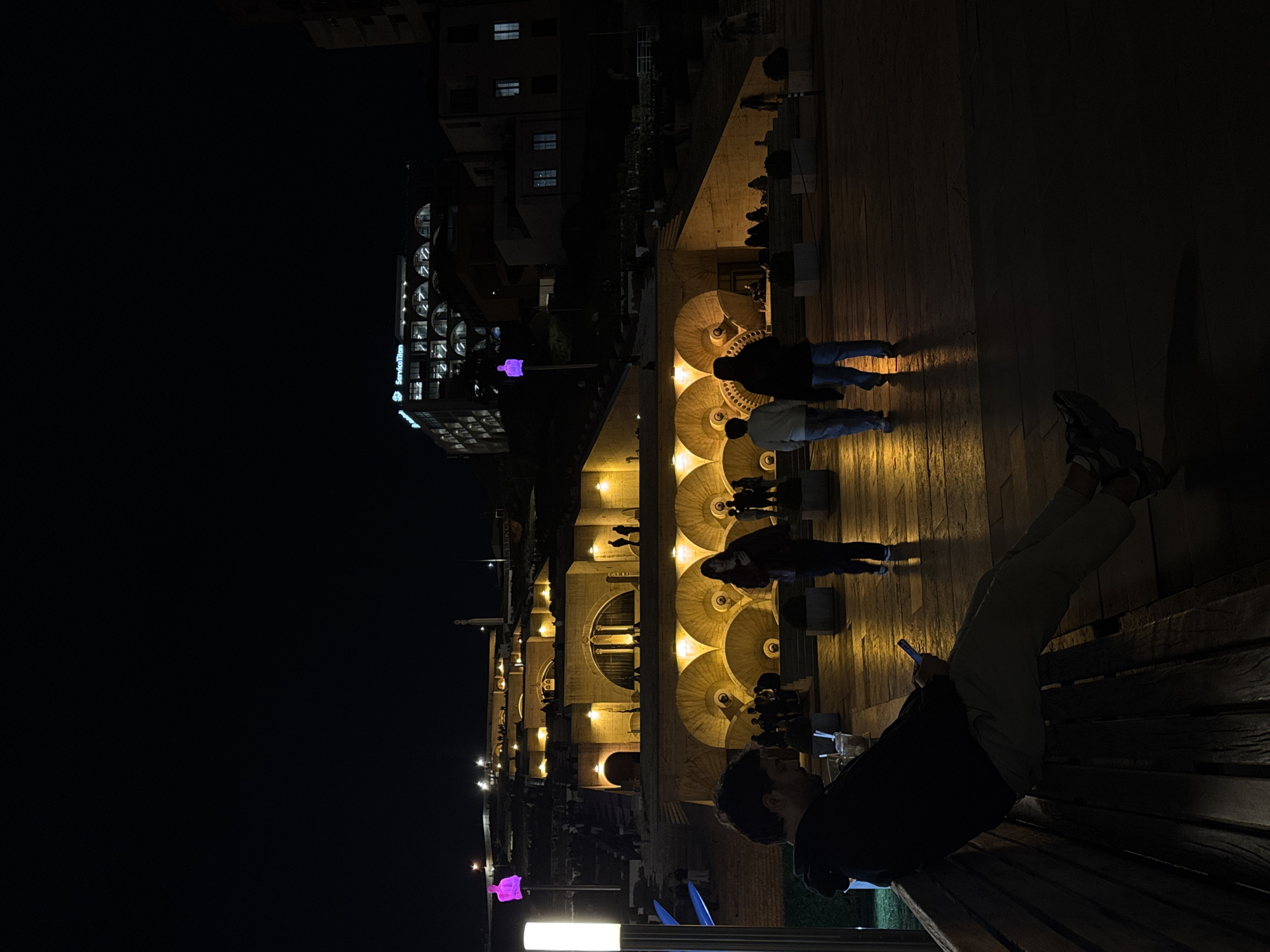 This night scene captures the illuminated Cascade Complex in Yerevan, Armenia, seen from an elevated perspective. The prominent multi-tiered architectural structure, characterized by its grand staircases and circular fountain basins, glows warmly under yellow artificial lights. These lights highlight the detailed stonework and steps, creating a striking contrast against the dark night sky.
In the lower right foreground, a person is seated on a wooden bench, leaning back comfortably. They are focused on a smartphone held in their hands, which emits a soft blue light. The individual wears light-colored pants and dark shoes.
Along the various levels of the Cascade, several other people are visible. Some appear as silhouettes walking or standing near the illuminated fountains, while others are slightly more discernible, exploring the complex. Two individuals, viewed from behind, stand prominently on one of the circular platforms in the mid-ground.
To the upper left, a modern building with distinctive circular windows is visible, featuring the illuminated blue text "ServiceTitan" on its side. Below this, near the Cascade, a glowing purple, abstract sculpture or light installation adds a modern artistic element to the environment. The clear night sky forms a dark backdrop, emphasizing the bright lights of the structures and the activity below. The scene conveys a calm, leisurely atmosphere as people enjoy the iconic landmark after dark.