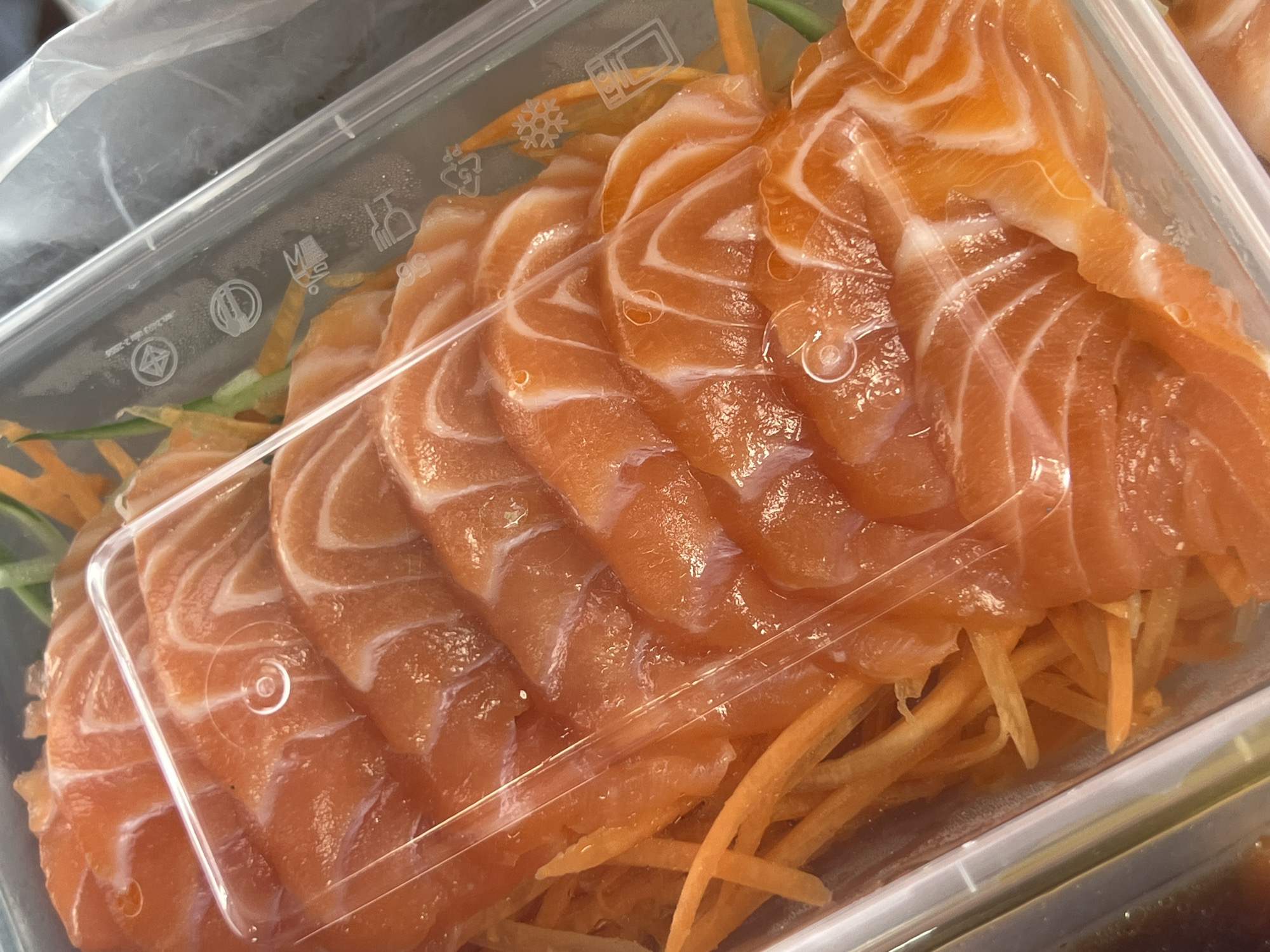 A close-up, overhead shot displays thinly sliced salmon sashimi arranged in a clear plastic container. The salmon pieces, a vibrant orange with distinct white marbling, are fanned out over a bed of shredded carrots and green garnish, possibly cucumber. A transparent plastic lid partially covers the food. The container has some embossed recycling symbols and text, including "PP" and what appears to be a "fork and knife" symbol. The lighting suggests it could be indoors or outdoors during the day, with reflections on the plastic lid and some condensation visible. The overall impression is of a fresh, prepared meal, likely for takeout or individual serving. There are no people visible, and the focus is entirely on the food. The scene is static, showcasing the texture and colors of the salmon. The location context of Baan Mae Nam, Thailand, is provided but not visually evident in the image itself.