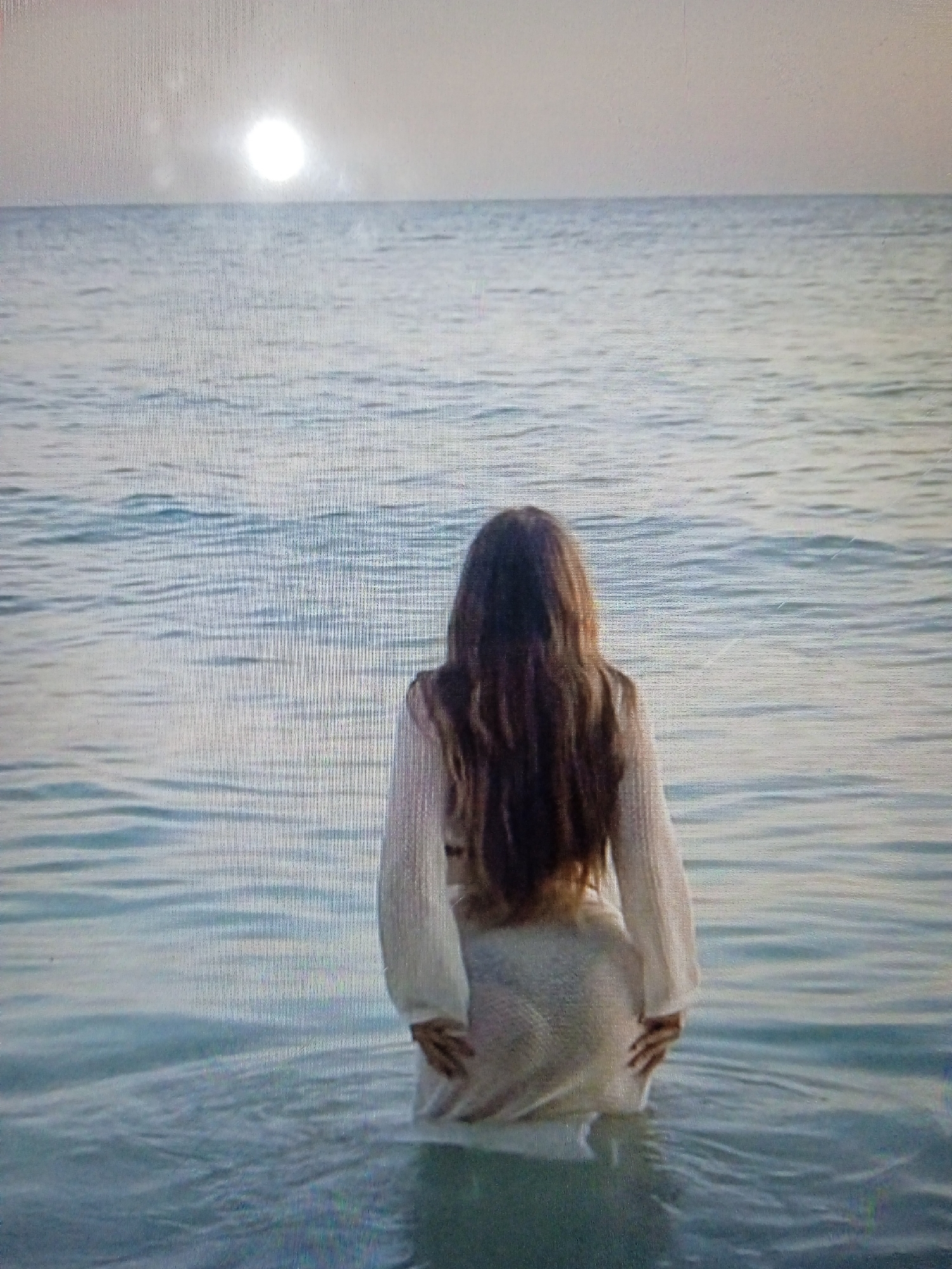 The image shows a person with long, dark hair standing in shallow water, facing away from the camera. The person is wearing a light-colored dress with long sleeves. The water appears calm with small ripples. In the background, the sun is visible as a bright orb near the horizon, illuminating a vast expanse of water. The overall scene suggests a serene, outdoor environment, possibly a beach or a lake.
There is no discernible information within the image or the provided crops to indicate the location as Chernivtsi, Ukraine. The scene is generic and could be any body of water with a clear horizon and sunlight.